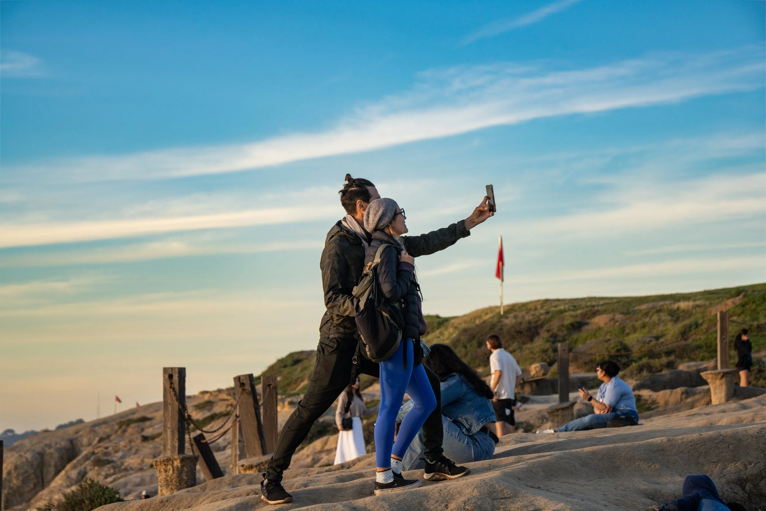 A couple taking a selfie on a rocky outdoor area during sunset with other people sitting and walking nearby.