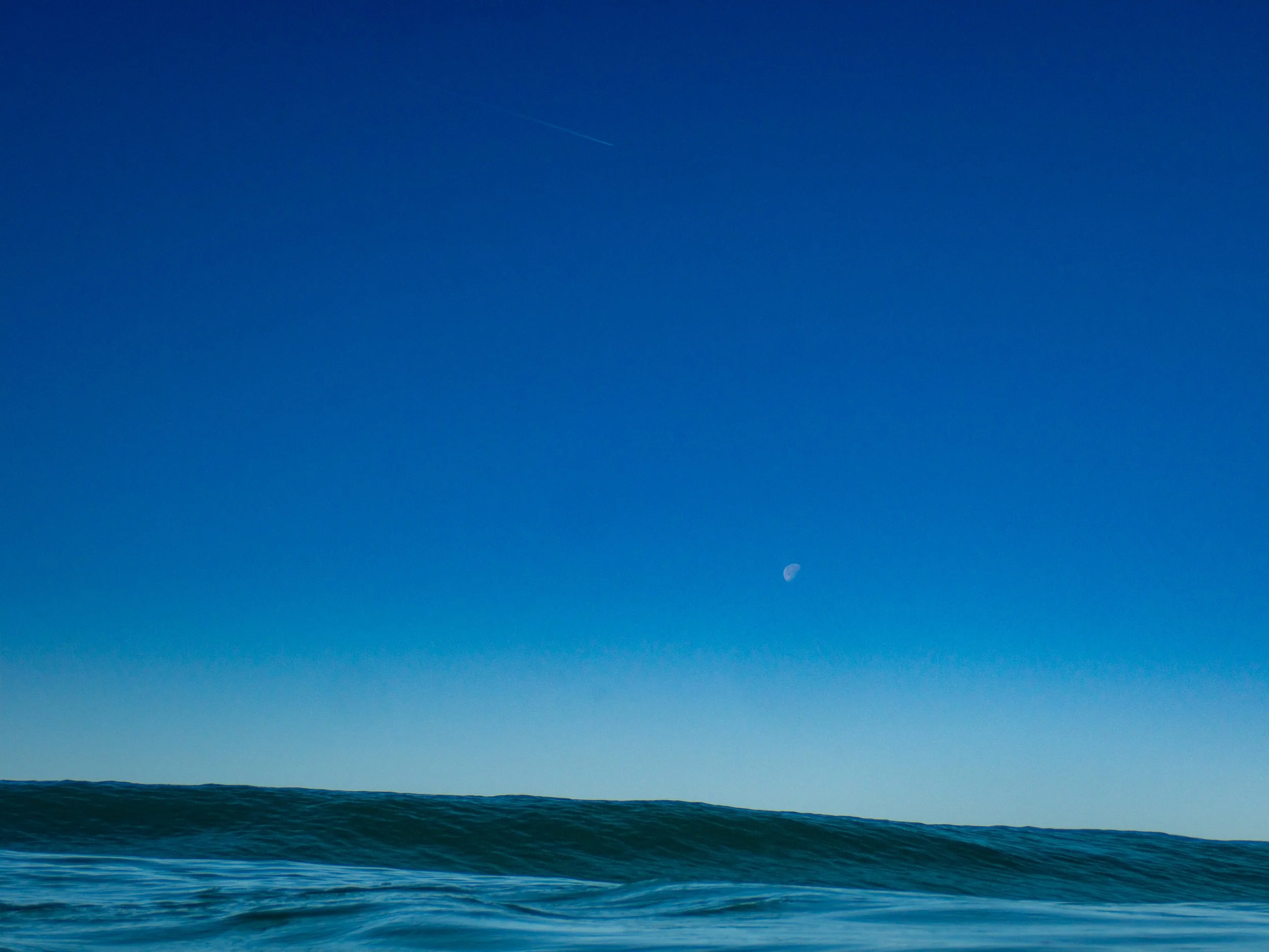 Ocean waves under a clear blue sky with a visible moon and a faint contrail.