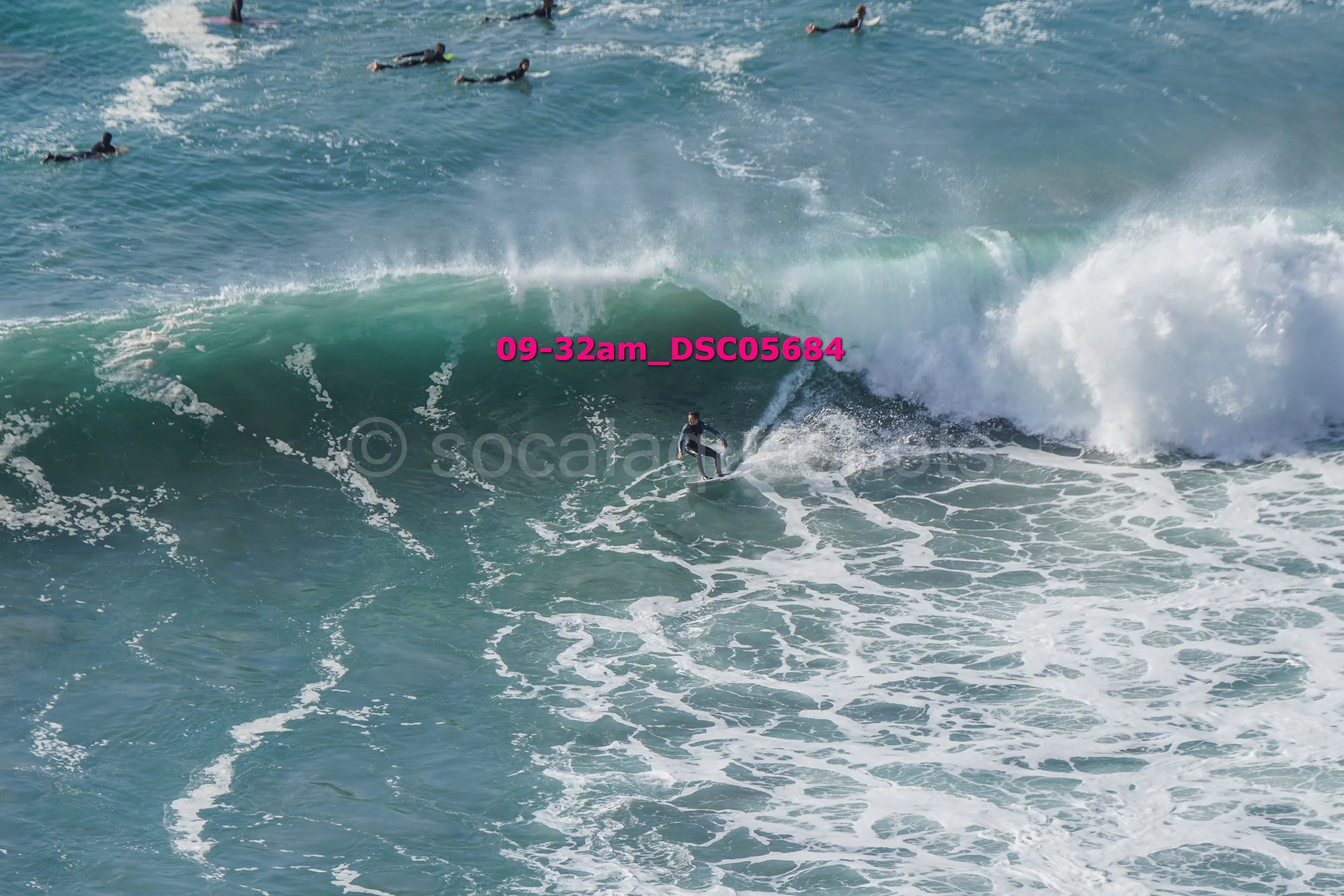 A person surfing on a large ocean wave, with several other surfers in the water in the background.
