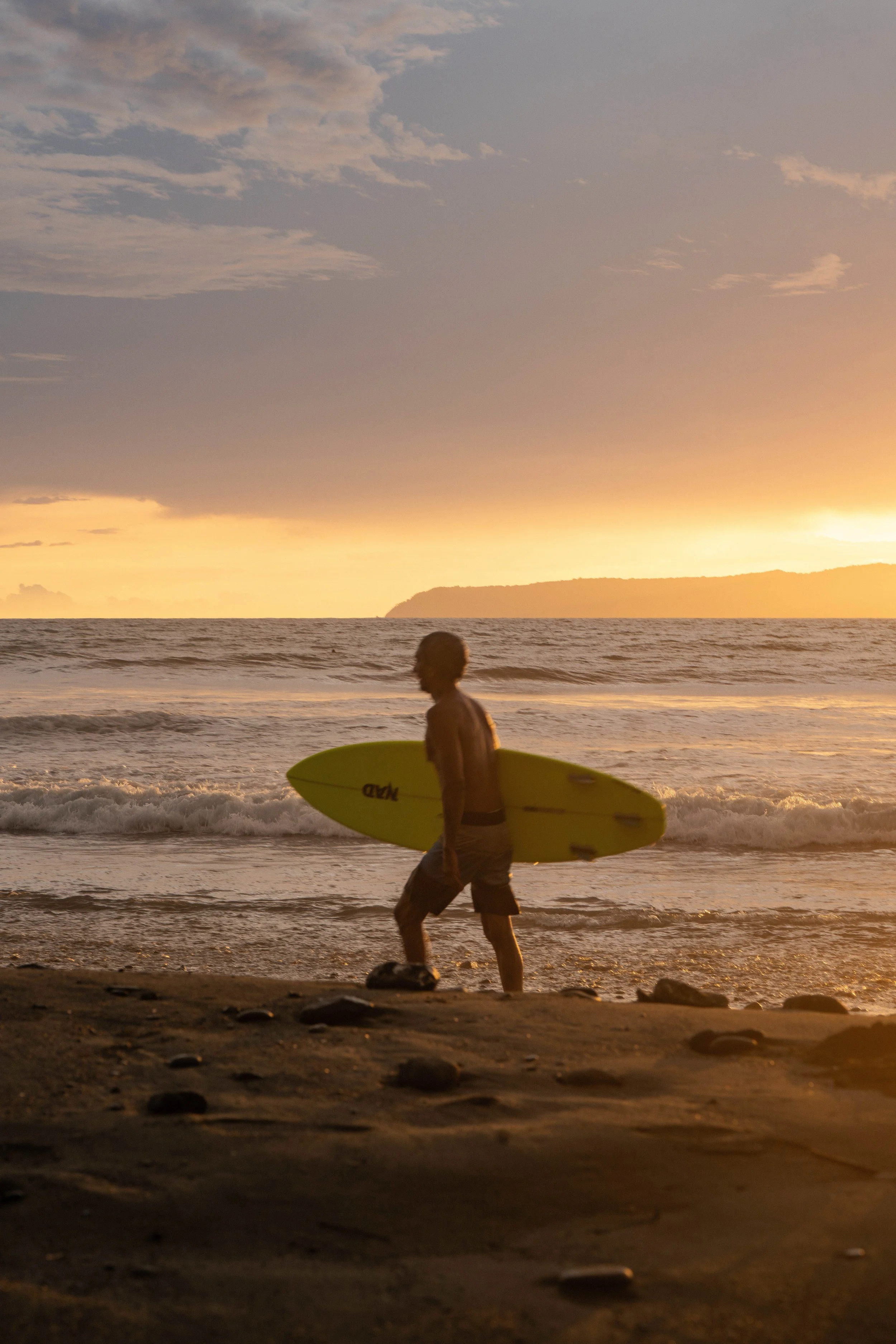 A person walking along the beach at sunset carrying a yellow surfboard, with the ocean and a distant landmass in the background.