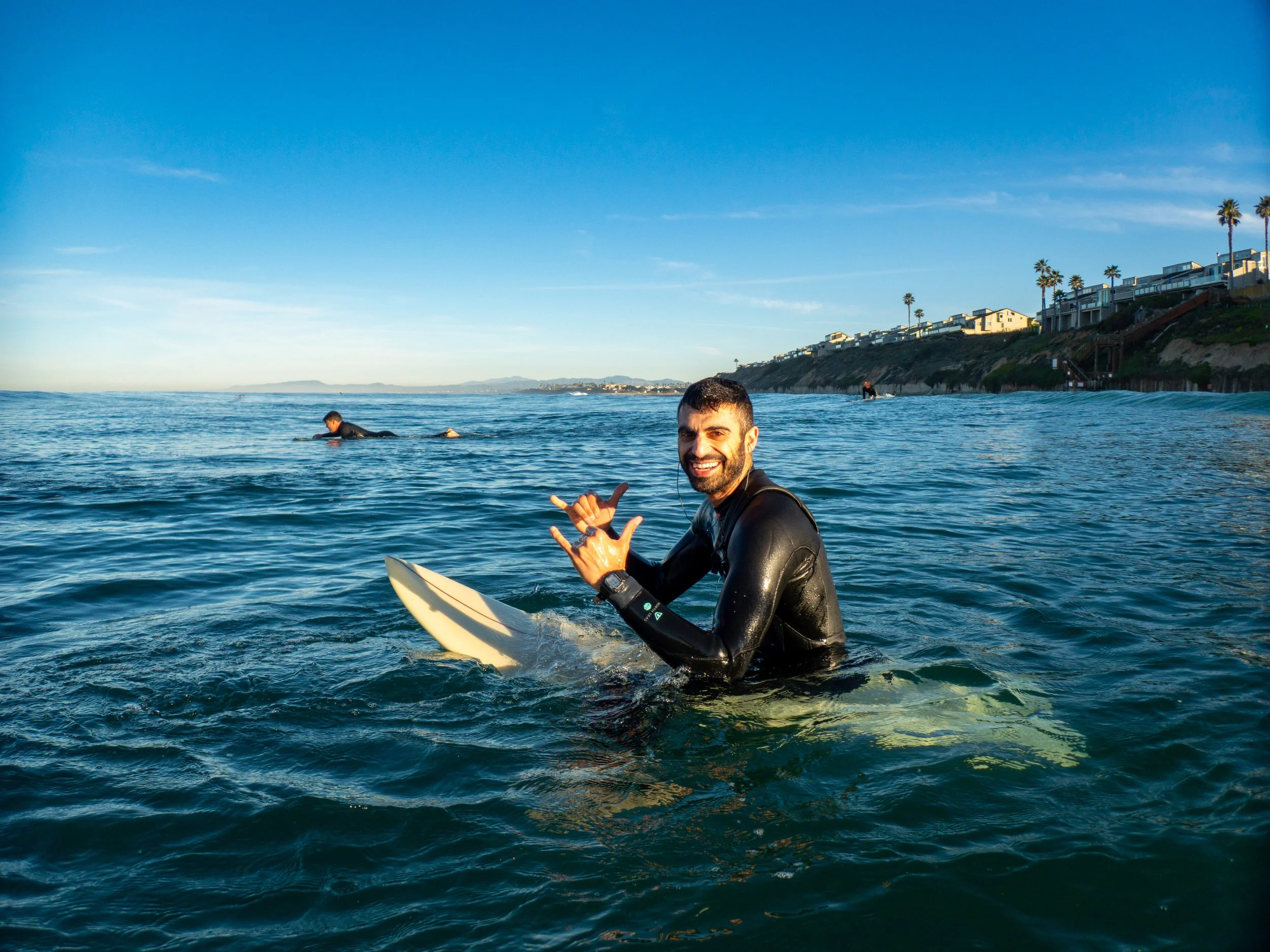 A man smiling and making a hand sign while sitting on a surfboard in the ocean with other surfers in the background and coastal houses and palm trees along a hillside under a clear blue sky.