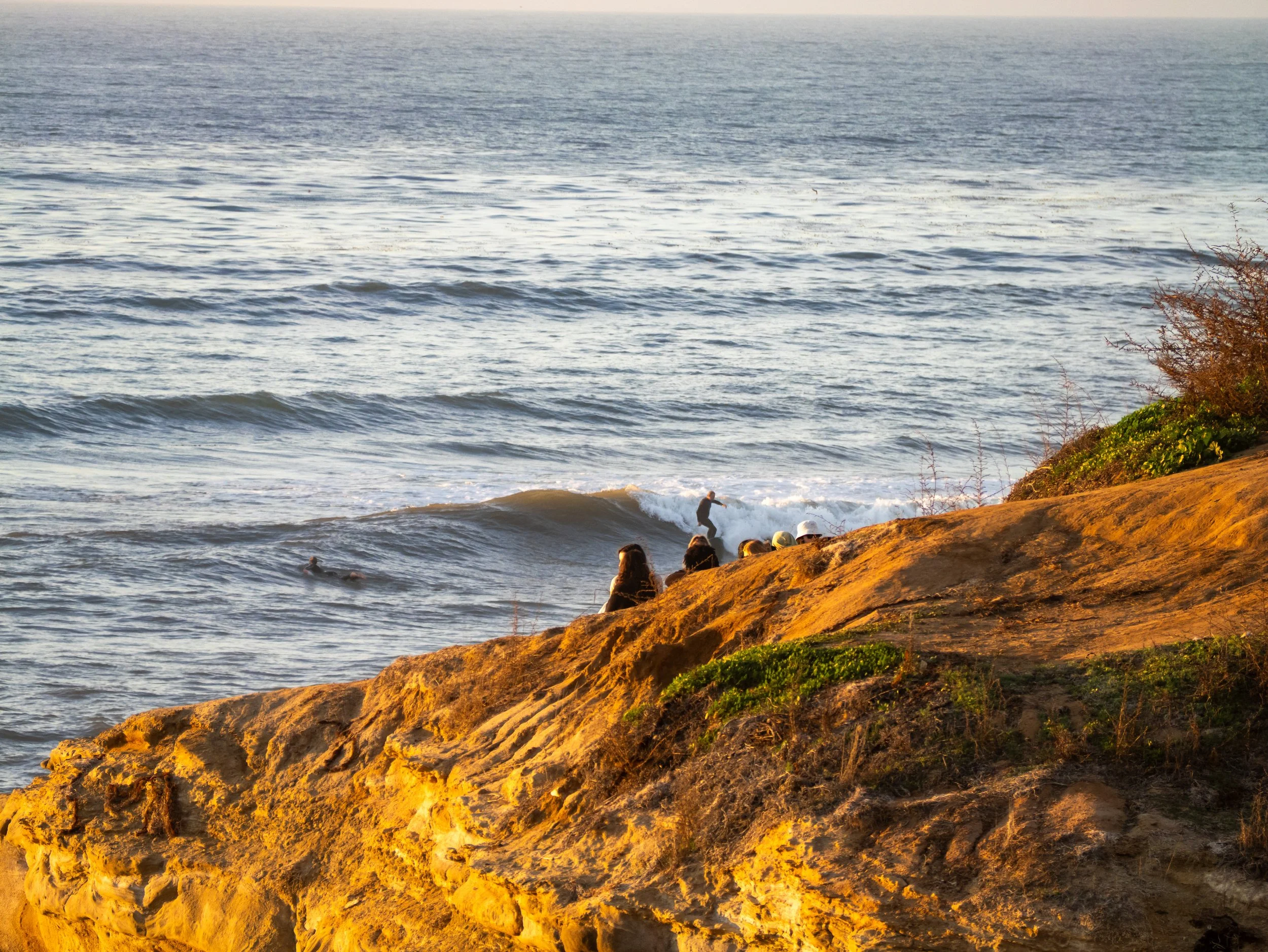 People sitting on a rocky cliff watching surfers in the ocean waves at sunset.
