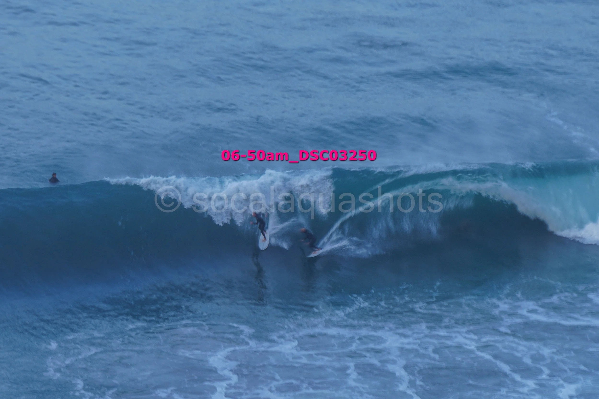 Two surfers riding a large wave in the ocean with a person floating nearby.