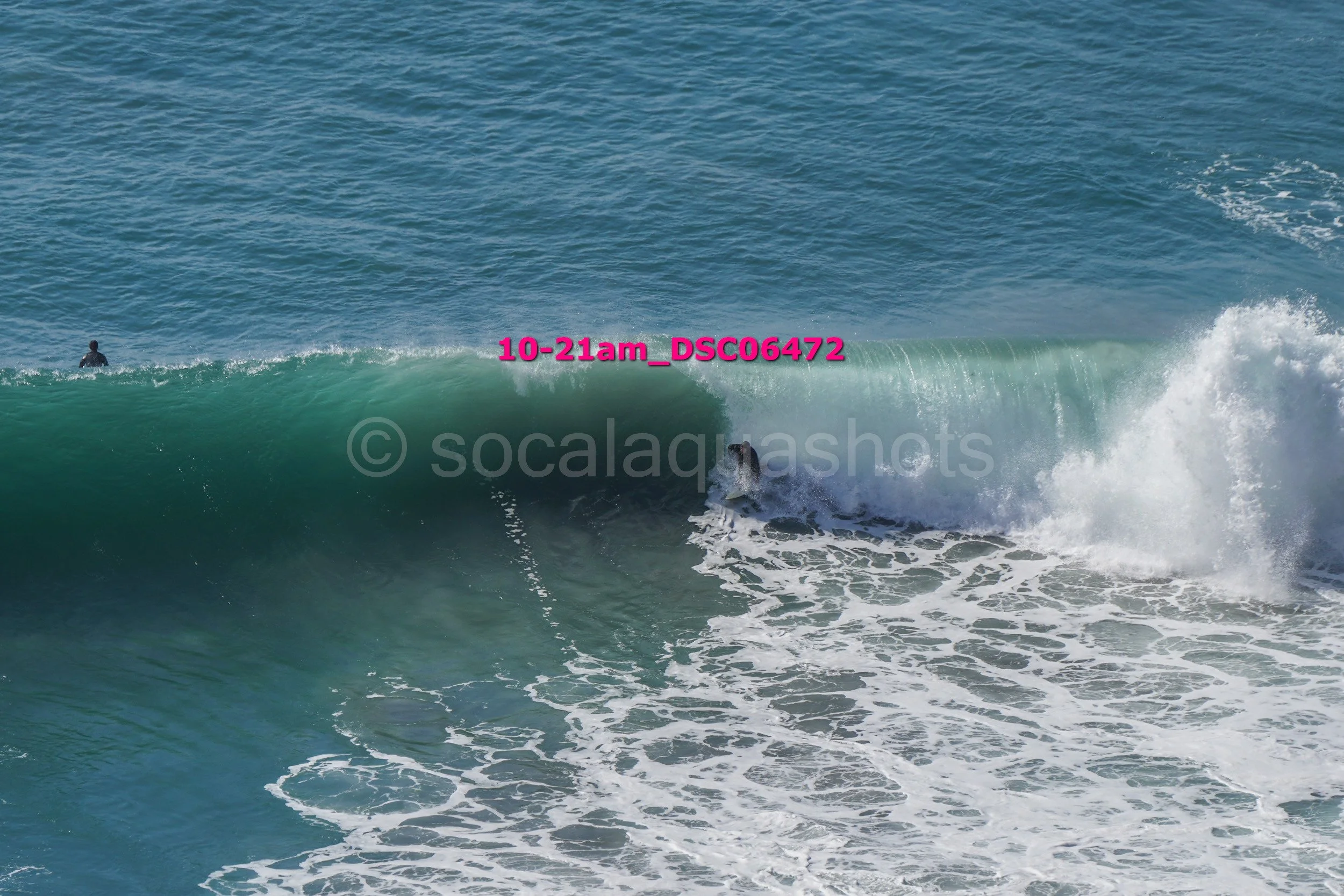 A person riding a surfboard on a large blue-green wave at the beach, with another person in the water nearby.