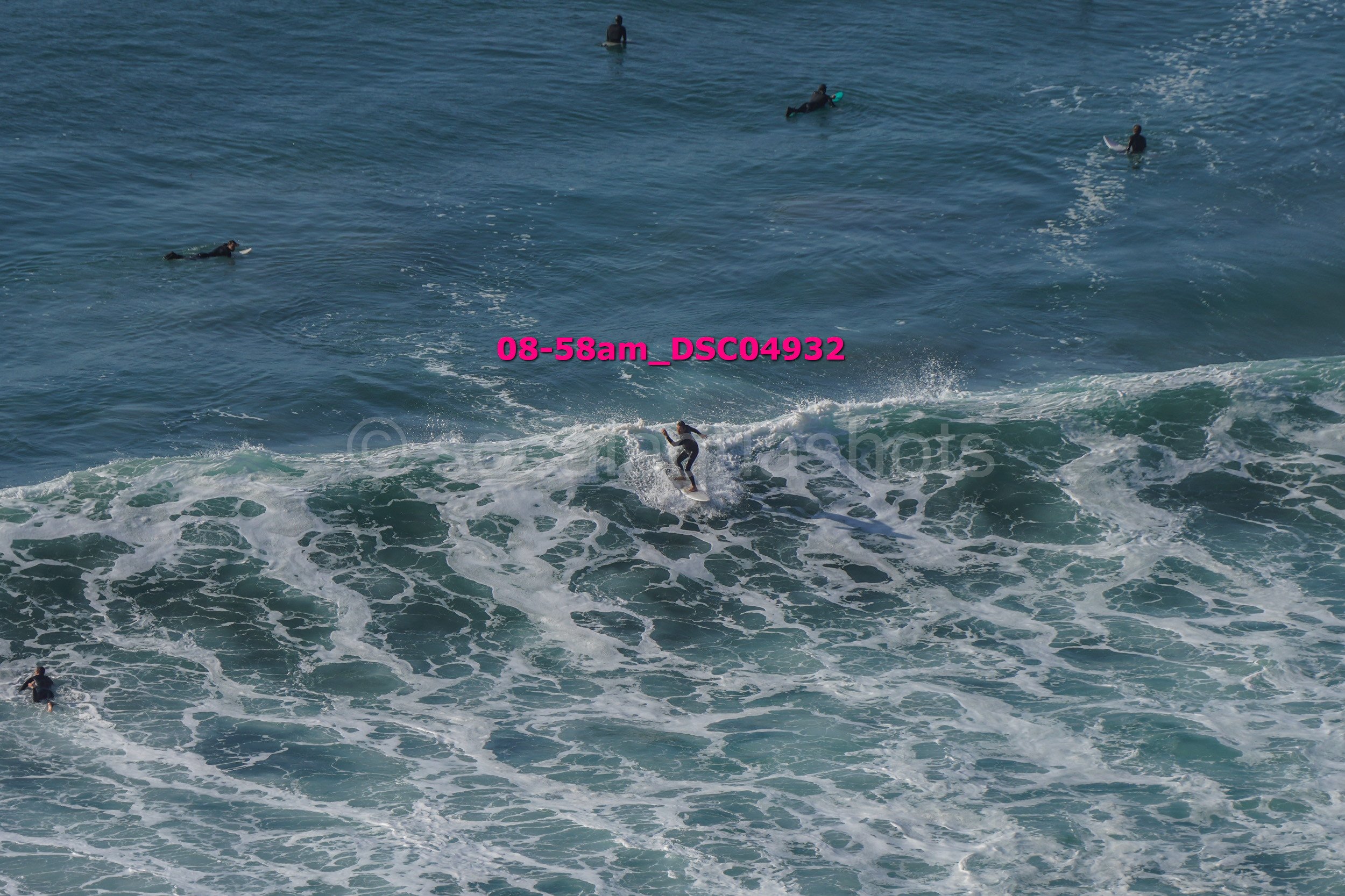 A person surfing on a wave in the ocean with several other people swimming or surfing in the water nearby.