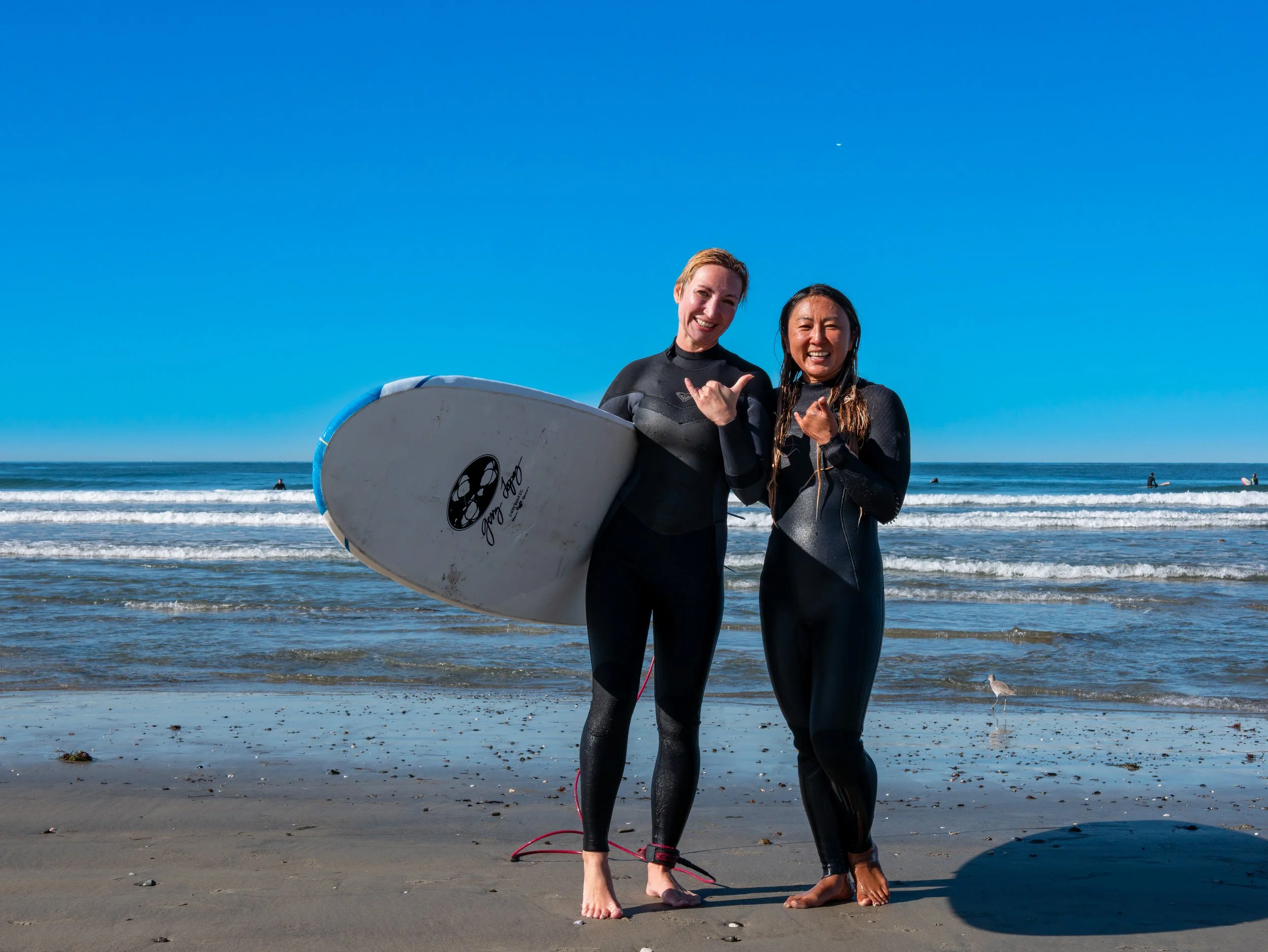 Two women in wetsuits standing on a beach, smiling and pointing at each other, with surfboards and the ocean in the background.