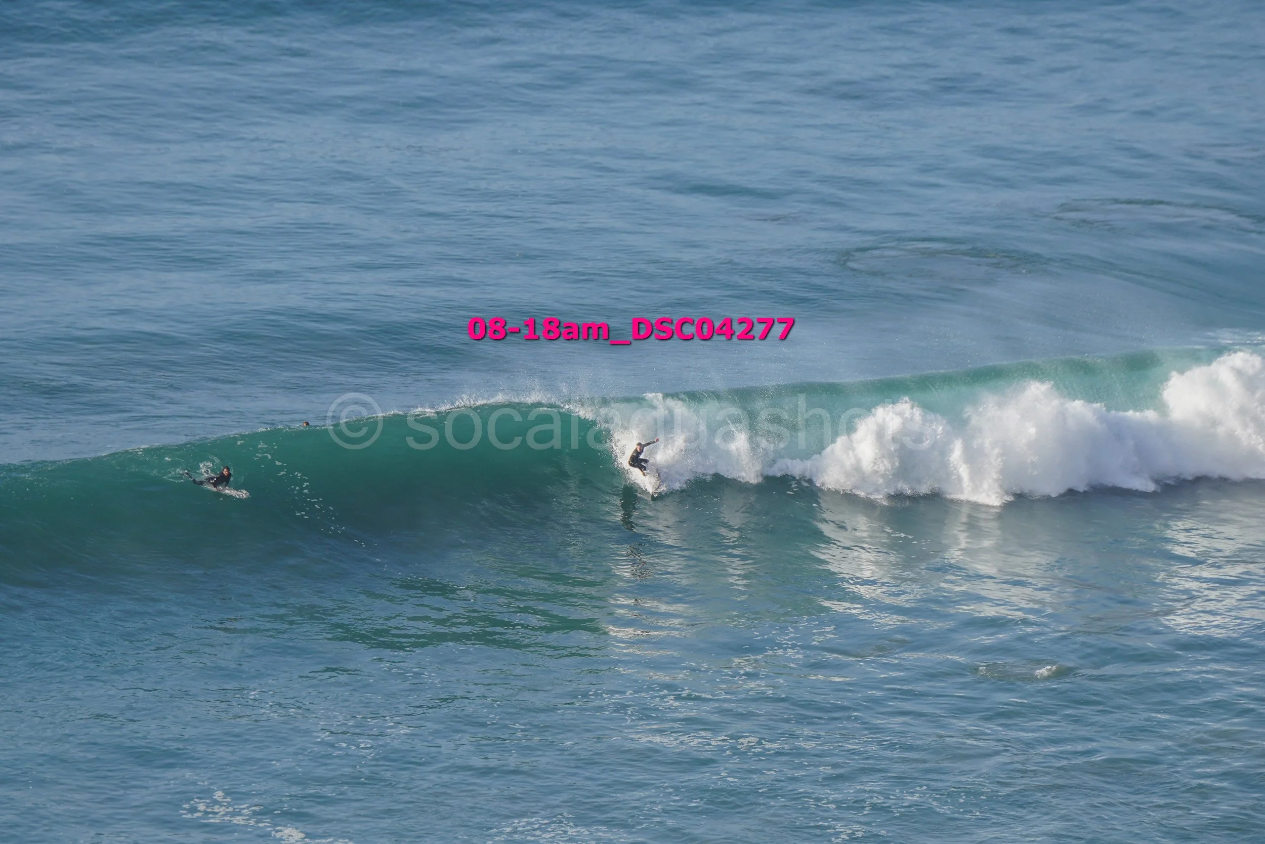 Two surfers riding a wave in the ocean with one surfer closer to the shore and the other further away; waves breaking in the background.