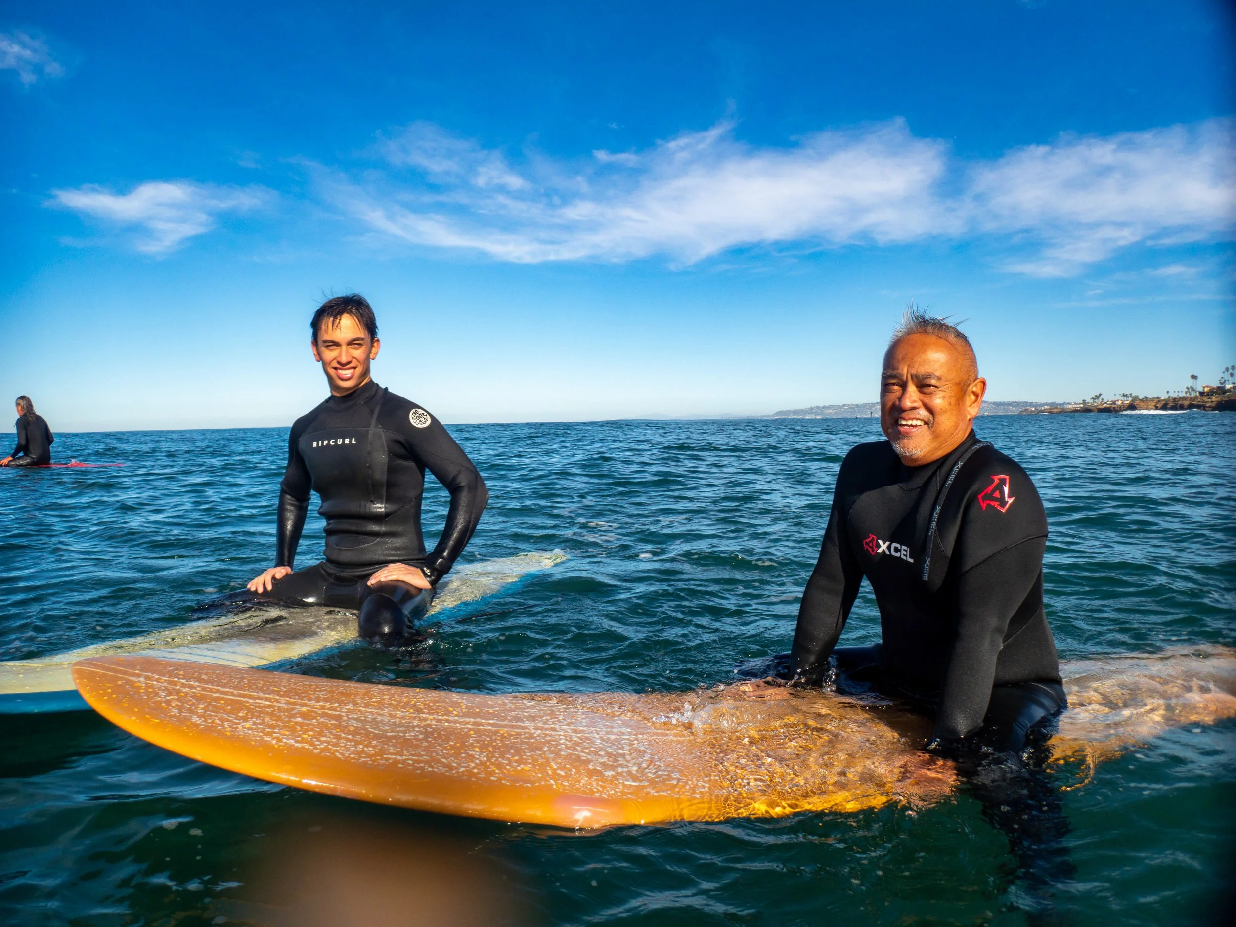 Two men in wetsuits on surfboards in the ocean, smiling at the camera with a clear blue sky and distant coastline in the background.