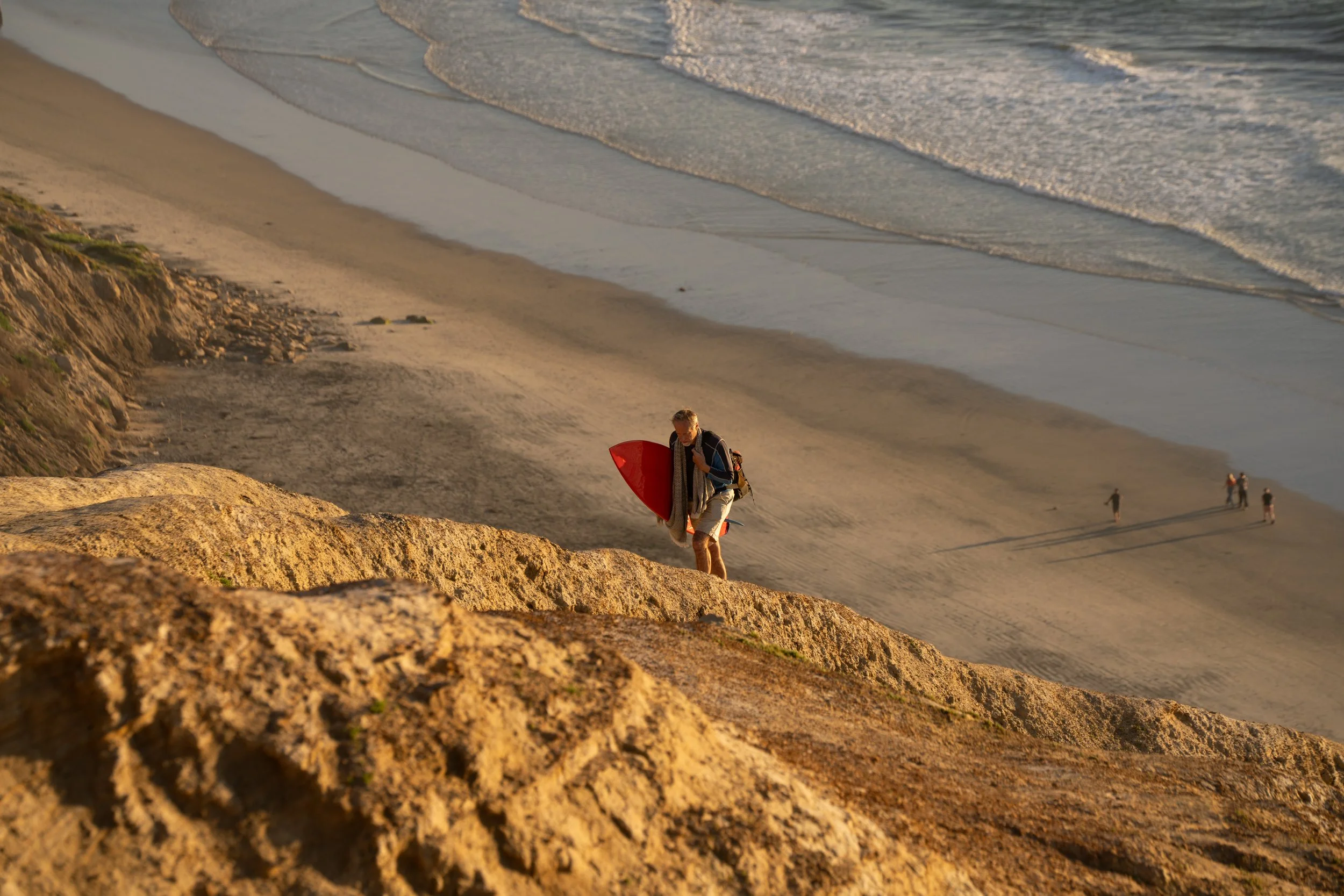 A man carrying a surfboard walking along rocks on a beach during sunset, with a few people visible walking on the sandy shore in the background.