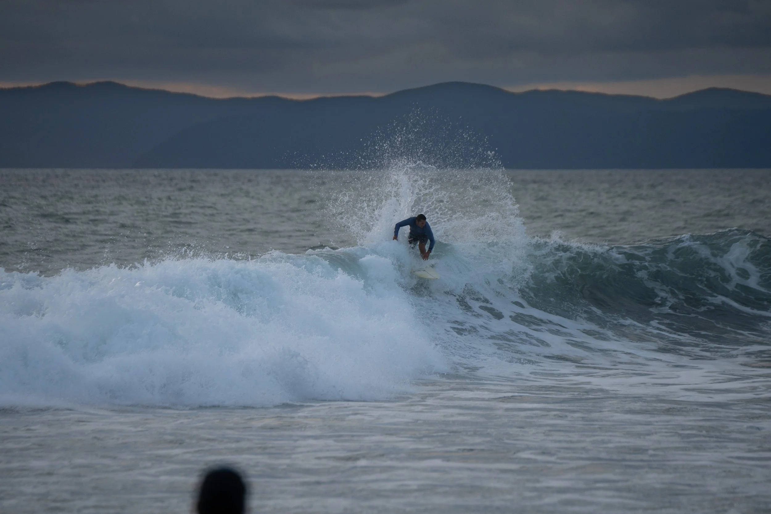 A person surfing on a wave in the ocean during cloudy weather, with mountains visible in the background.
