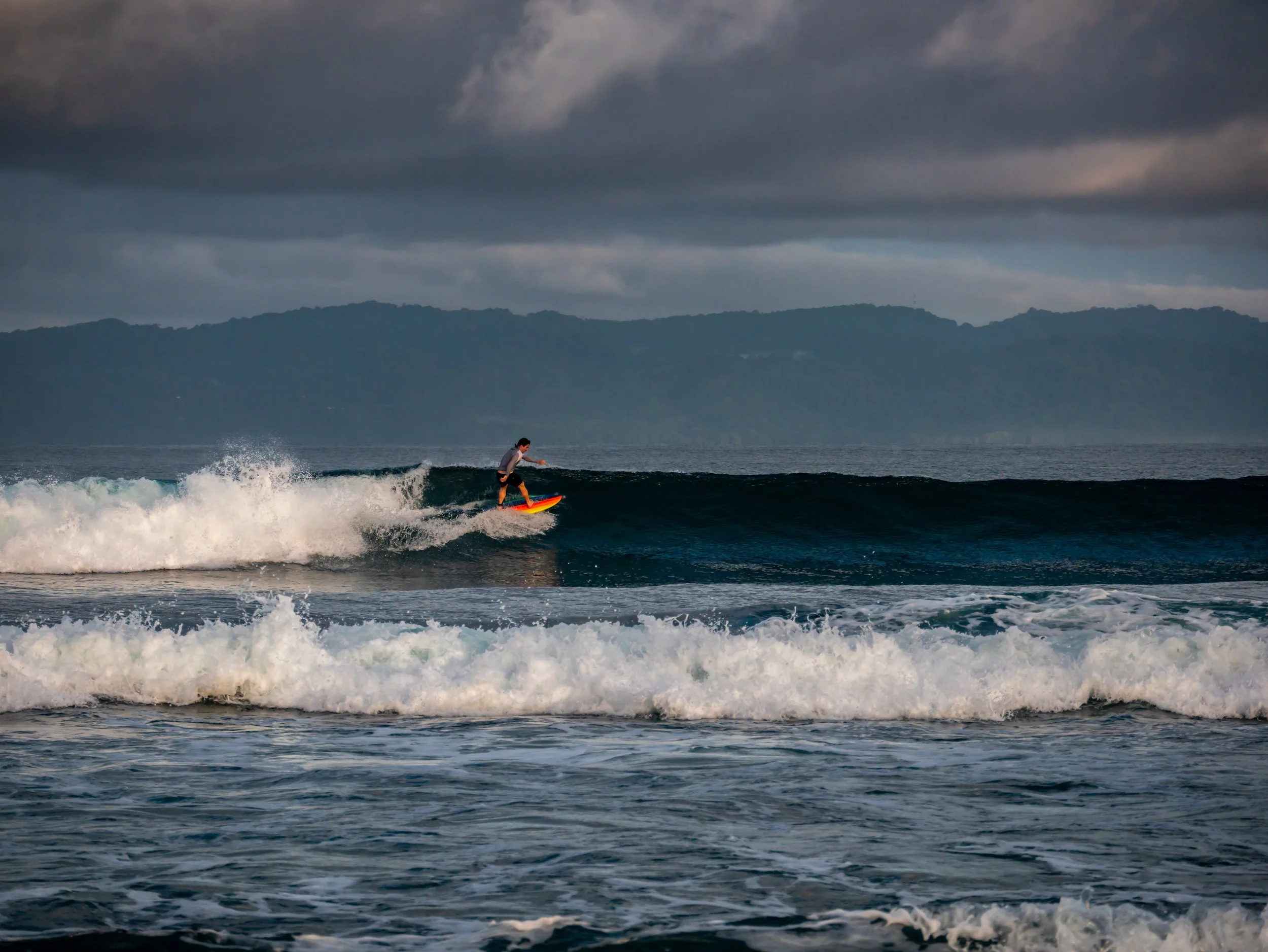 Surfer riding a wave on a cloudy day with hills in the background.