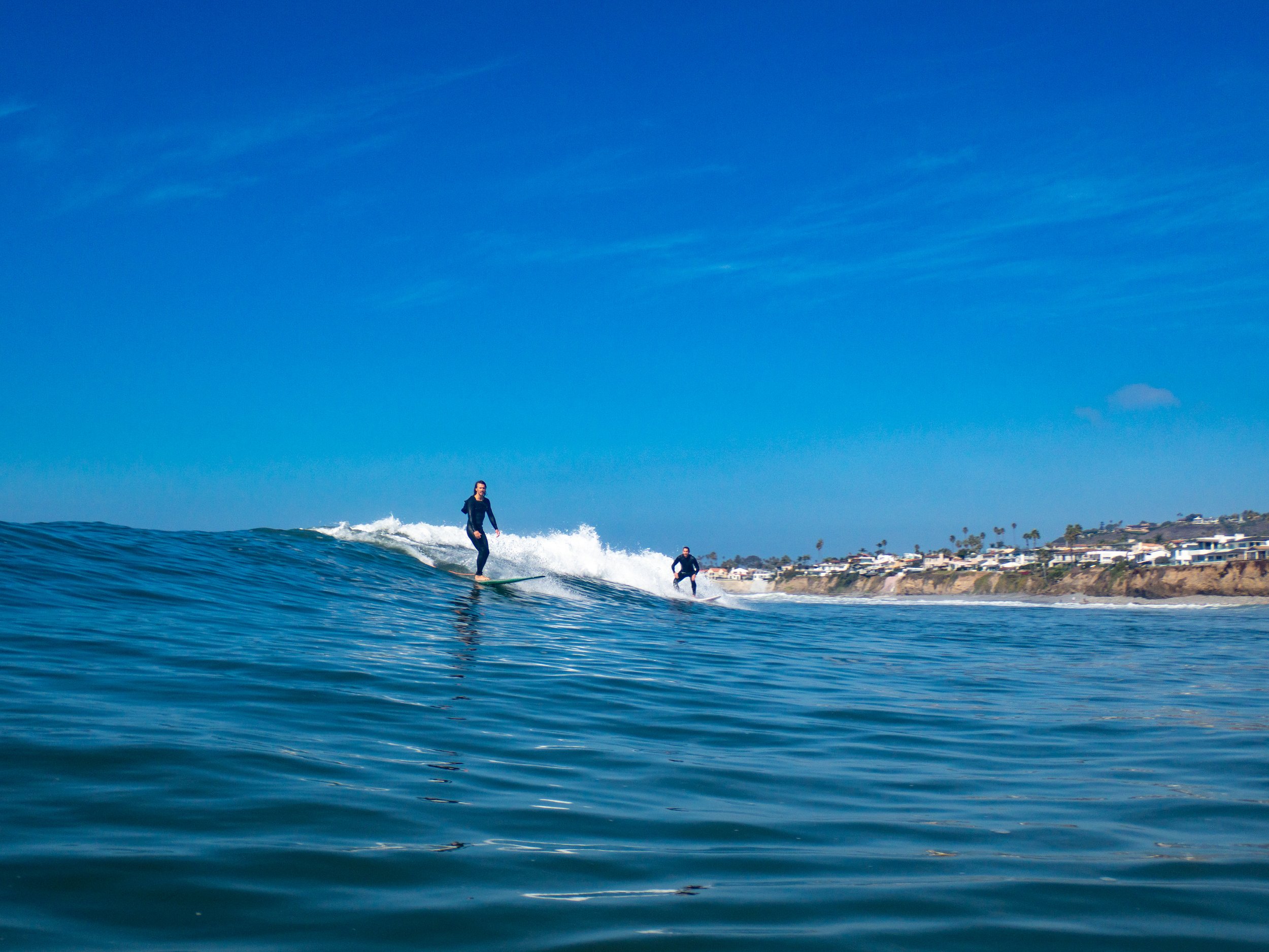 Two surfers riding on a wave near the coast under a clear blue sky, with buildings and palm trees on the shoreline in the background.