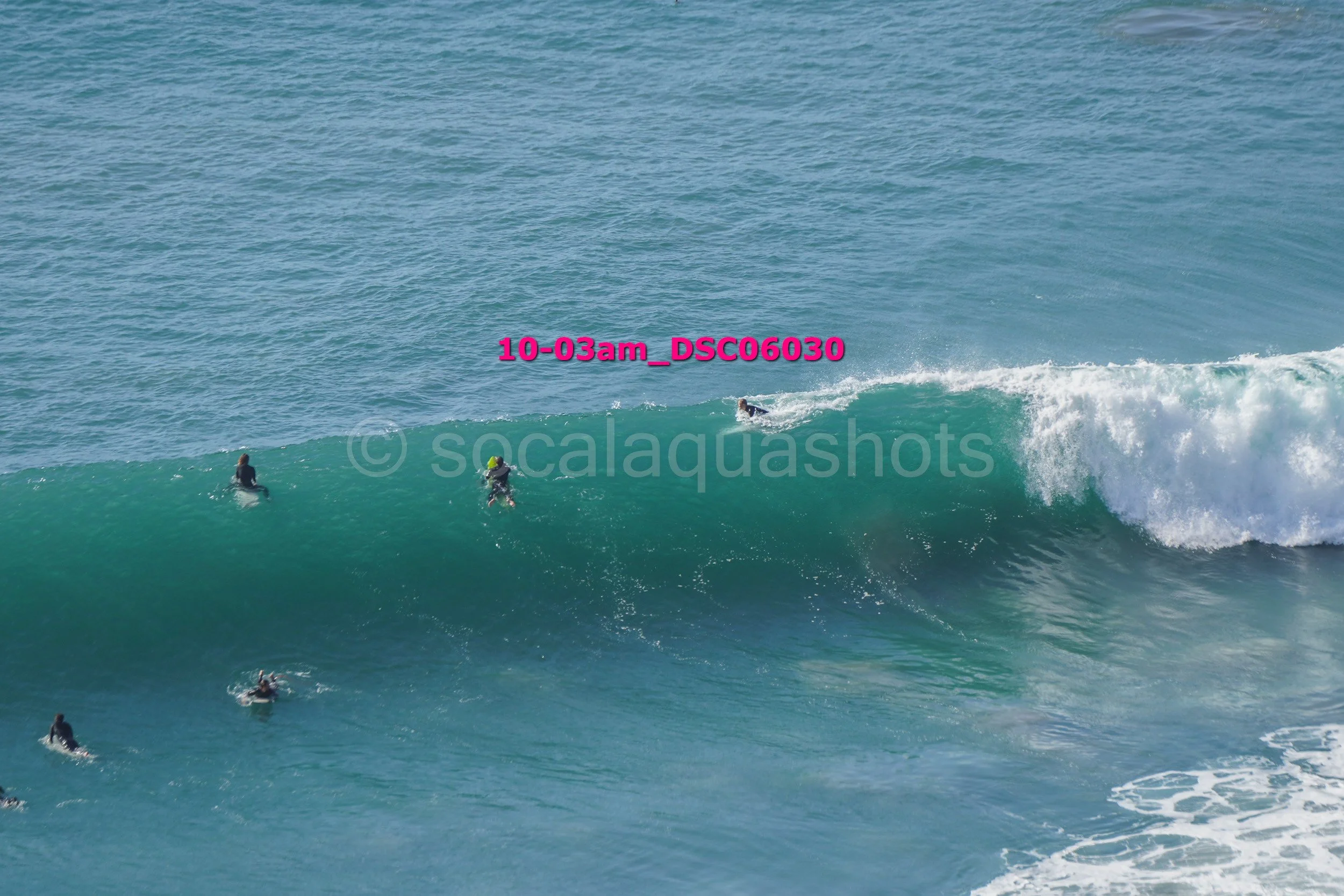Surfers riding a large ocean wave, with some surfers paddling in the water nearby.
