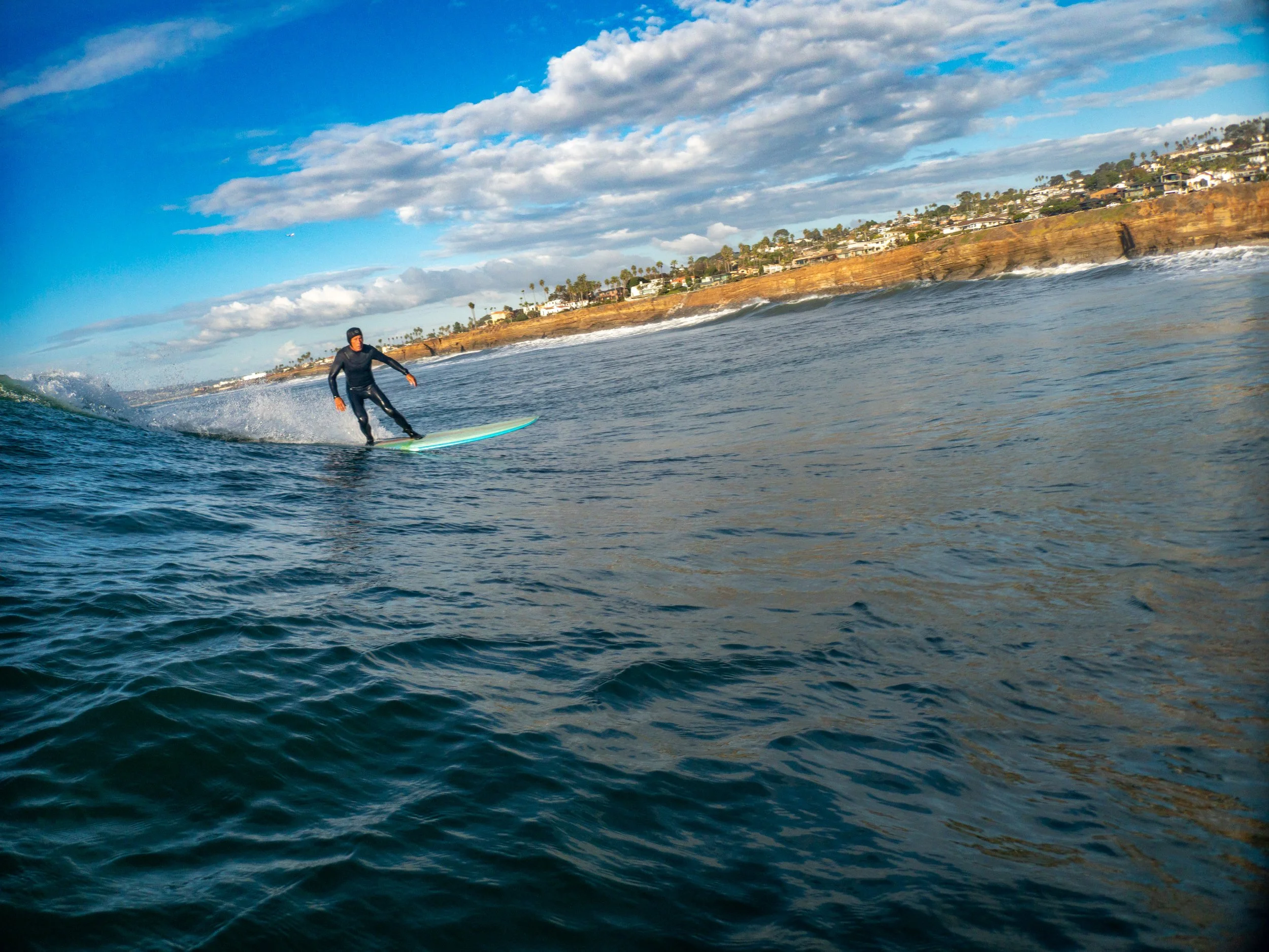 A person surfing on the ocean near a rocky coastline with houses on the cliffs and partly cloudy sky.