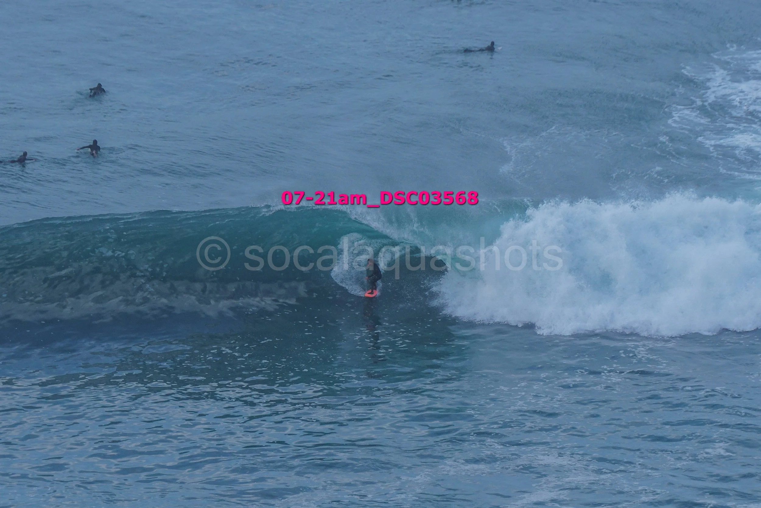 A surfer riding a wave at the beach with several other surfers in the water. The photo is taken at 7:21 am and includes a watermark from socalquash.