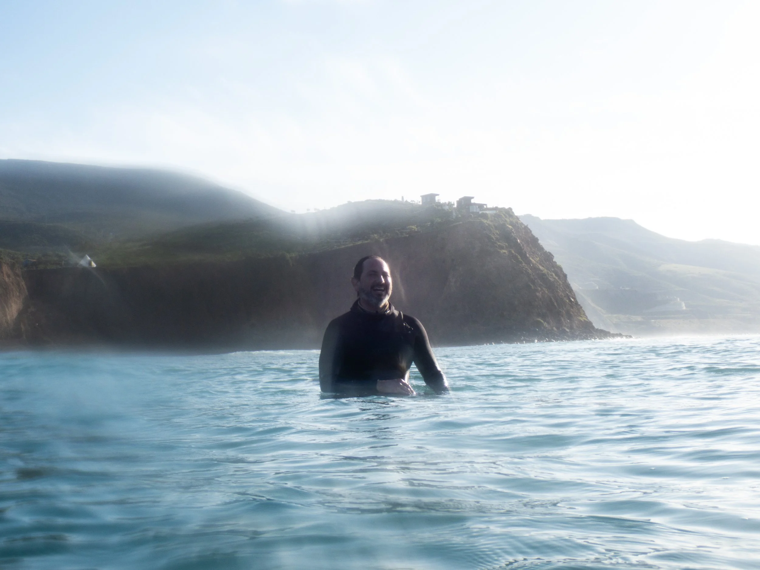 A man in a black wetsuit standing in the ocean with a misty coastline and hills in the background, smiling at sunset or sunrise.