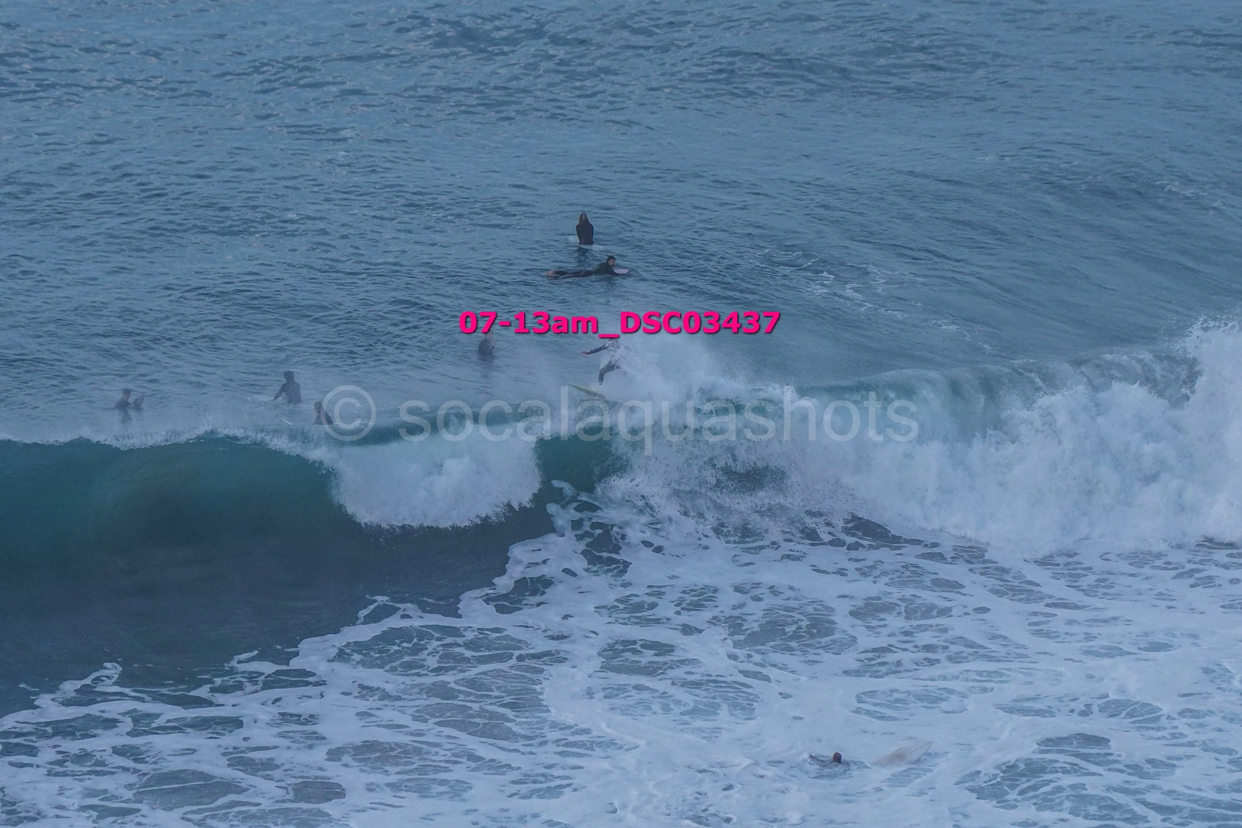 Several surfers in the ocean; some are riding a wave, others waiting in the water. The photo has a pink timestamp overlay '07-13am_DSC03437' and a watermark 'socalaquashots'.