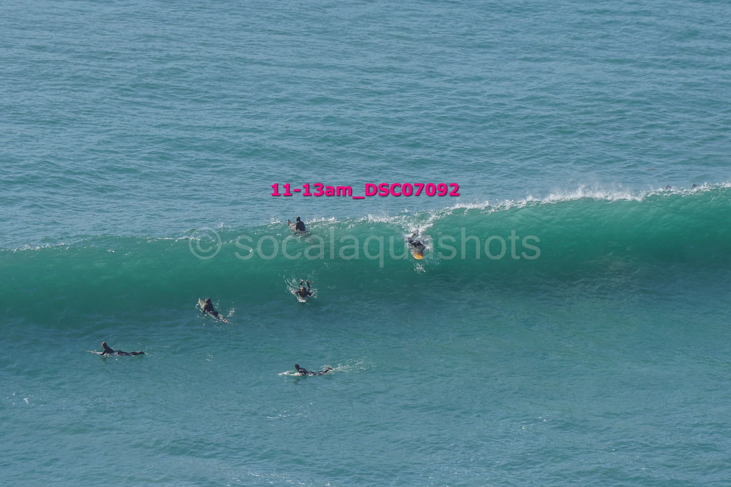 Group of surfers paddling and surfing on a large blue-green wave in the ocean.