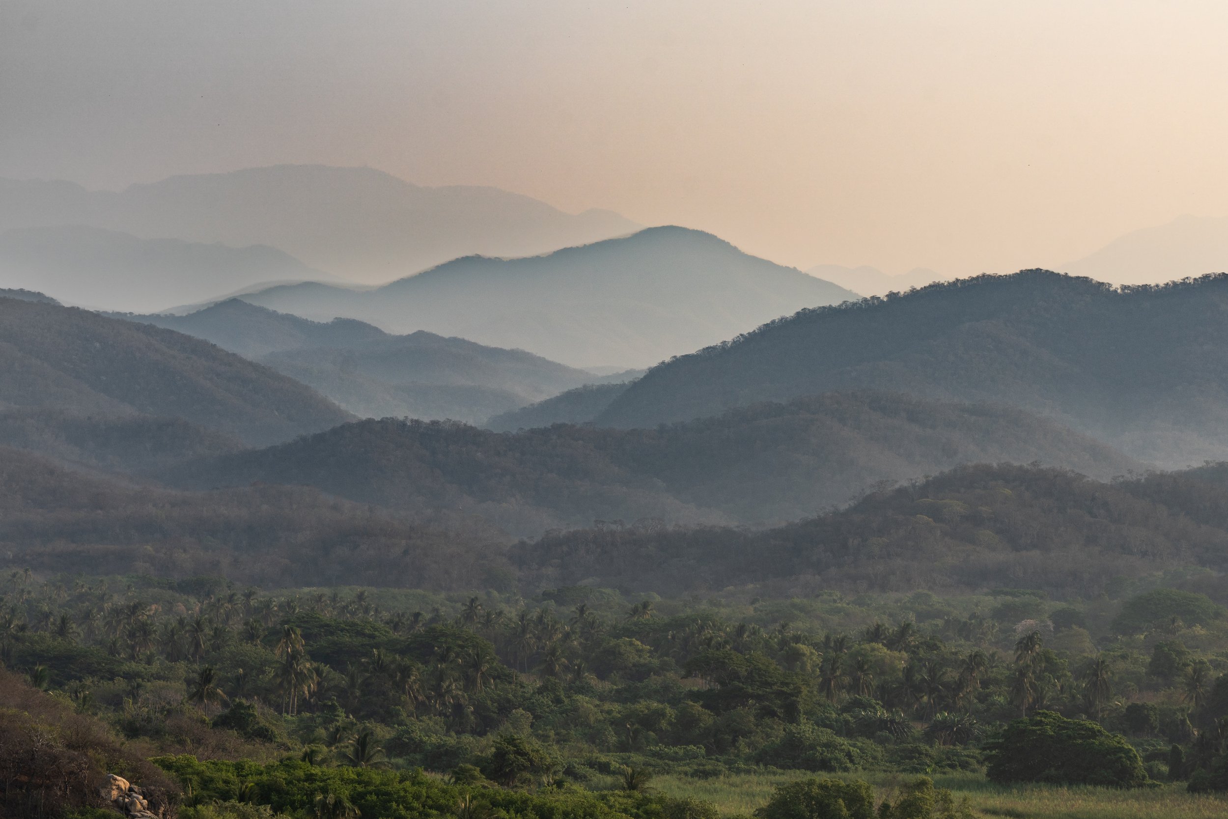 A layered landscape of rolling hills and mountains covered with greenery, fading into the distance with a hazy sky.