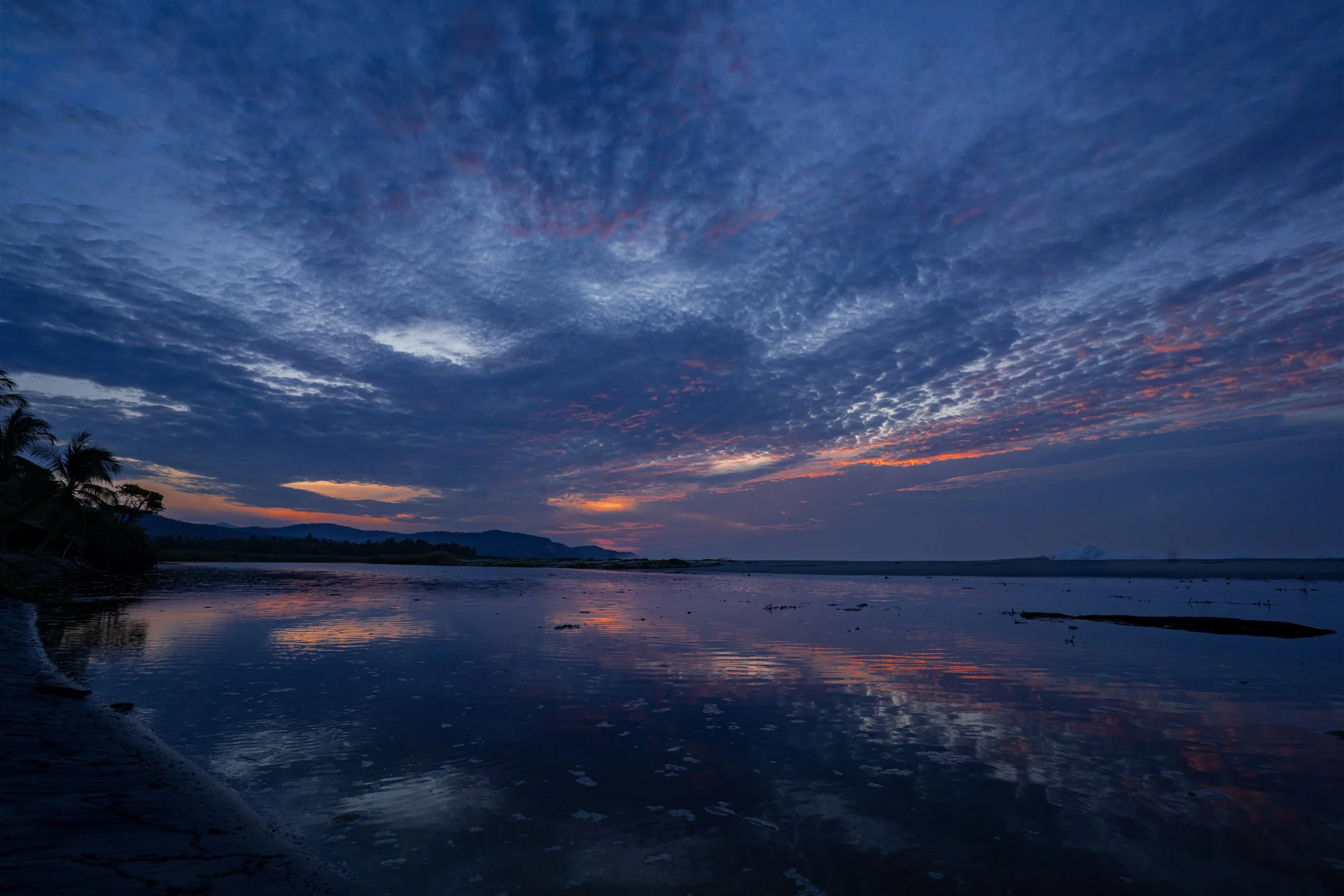 A scenic view of a sunset over a river with clouds in the sky and mountains in the background.