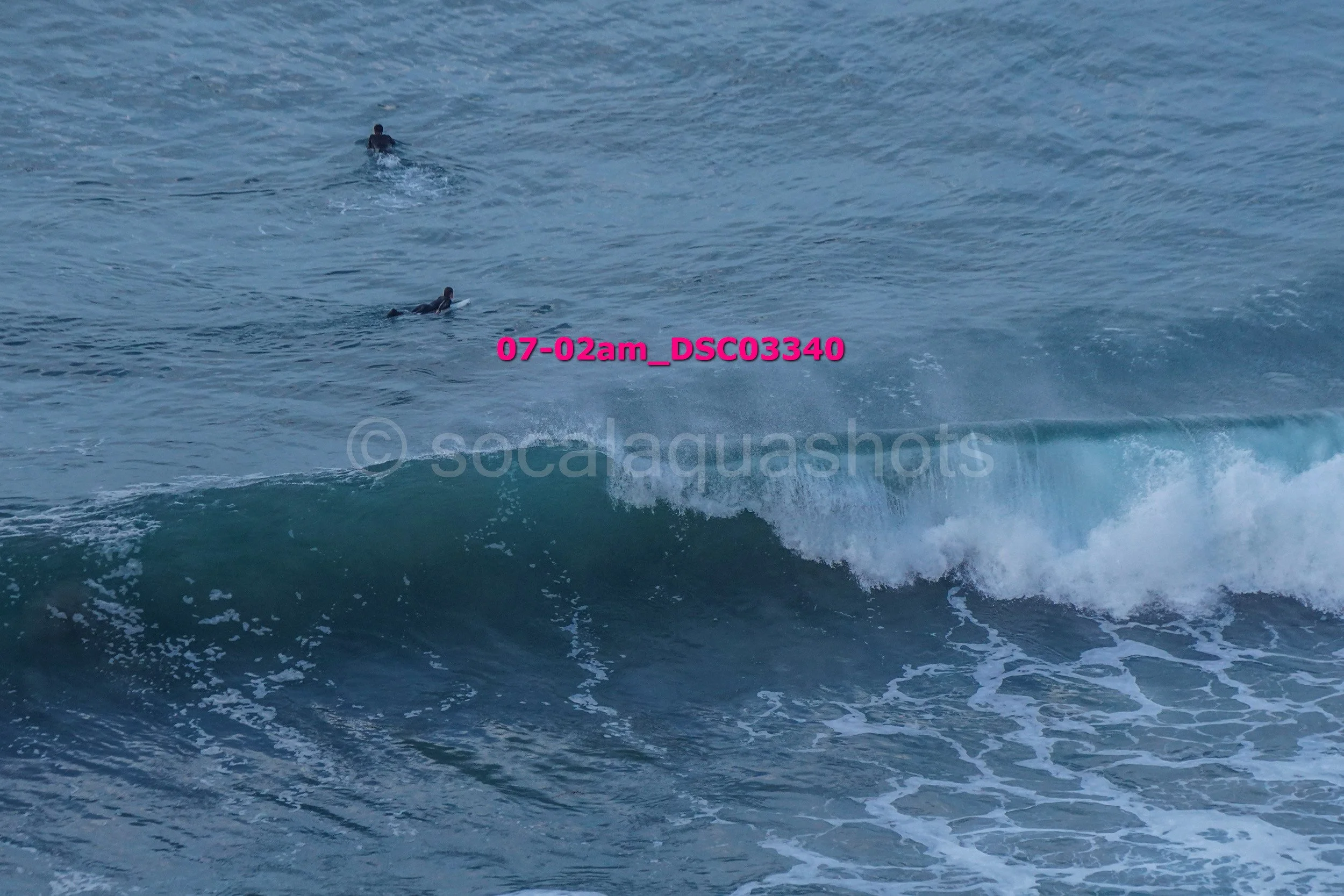 Two surfers in the ocean, one sitting on a surfboard and the other paddling on their surfboard near a wave.