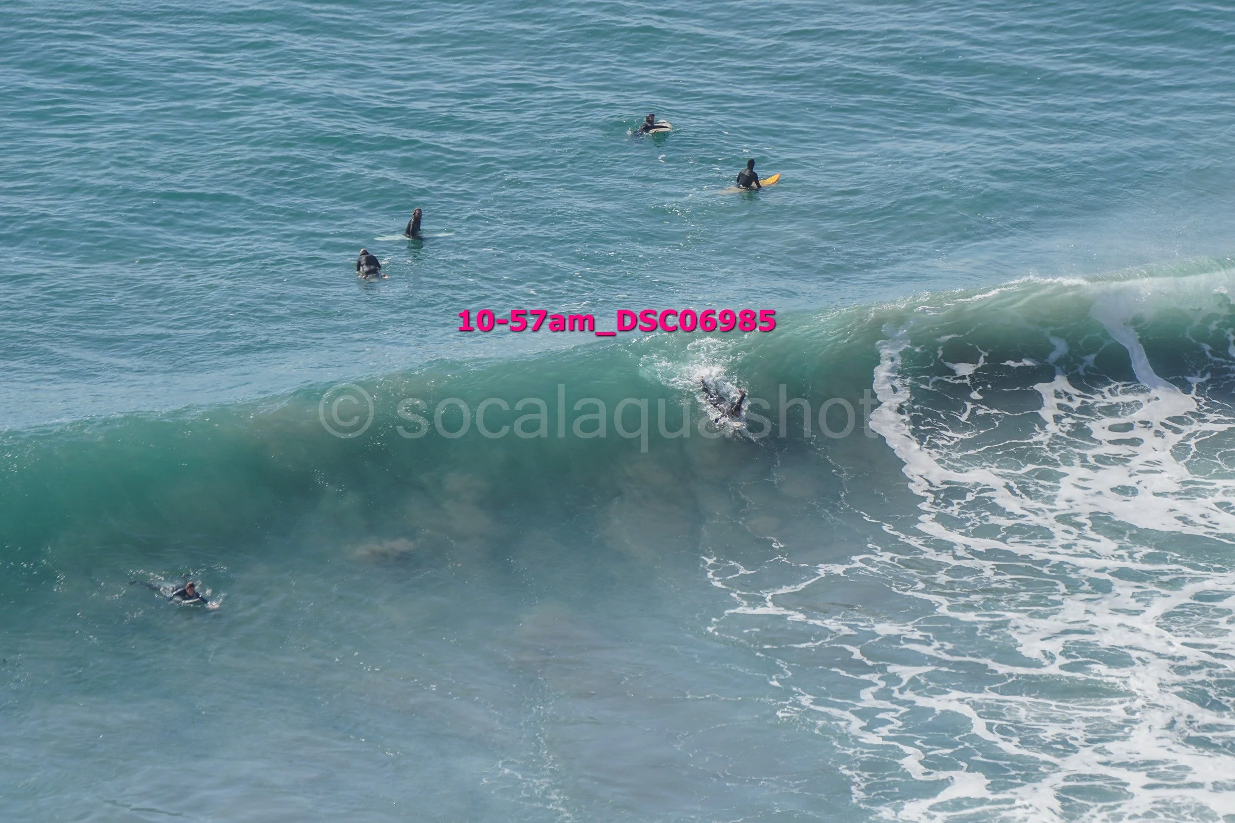 Surfer riding a large wave with several people on surfboards in the water nearby.