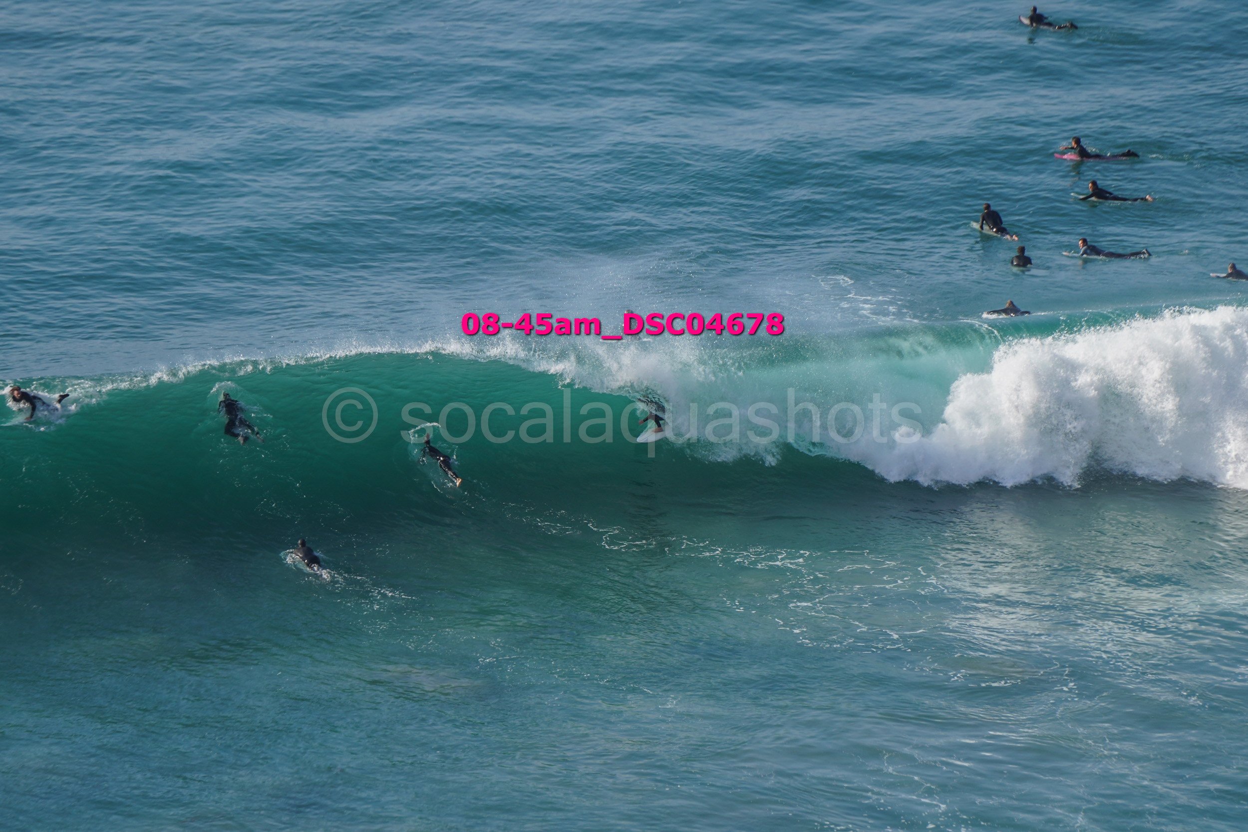 Multiple surfers riding and waiting for waves in the ocean with a prominent pink timestamp and watermark overlay.