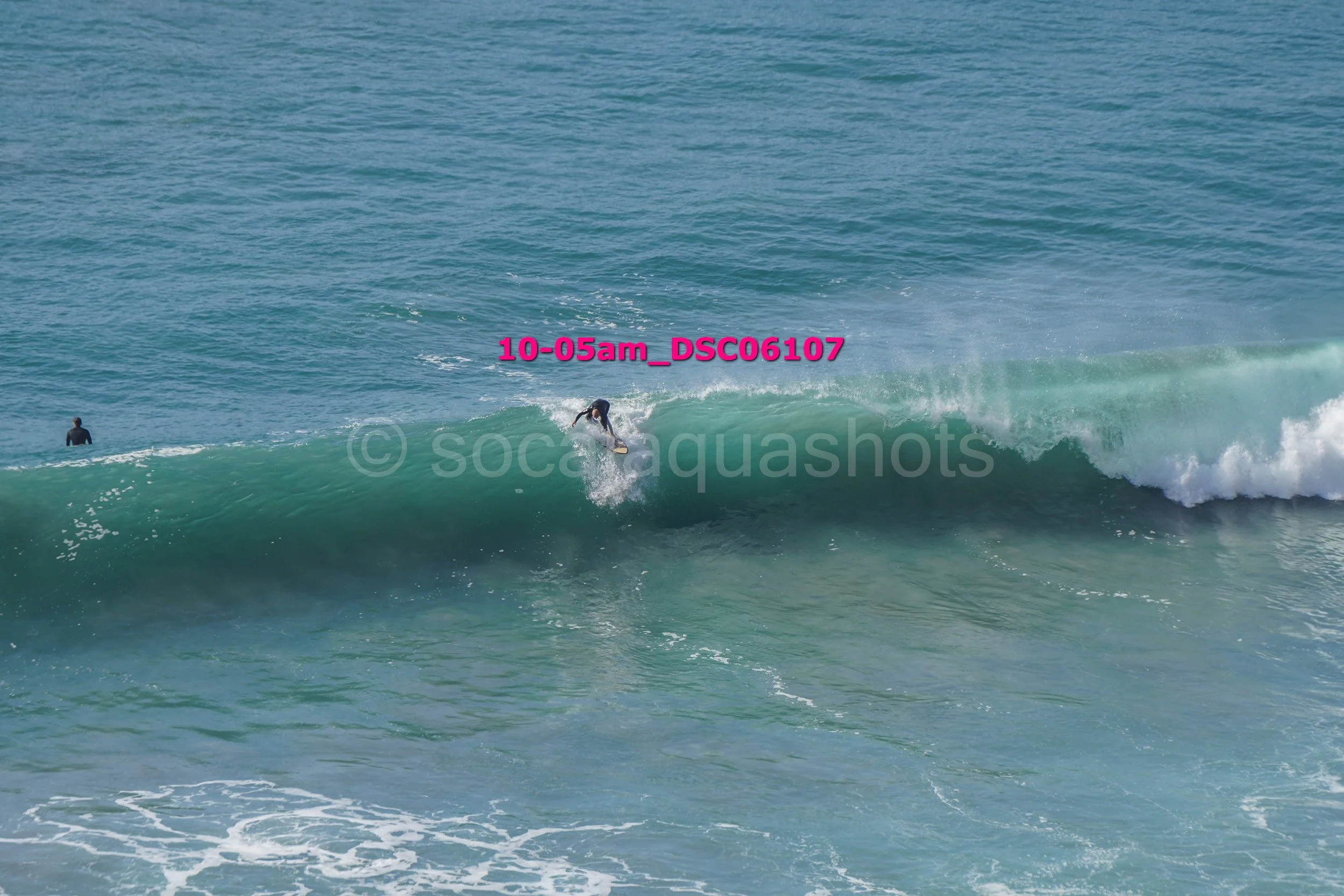 Person surfing on a wave in the ocean with another person standing in the water nearby.