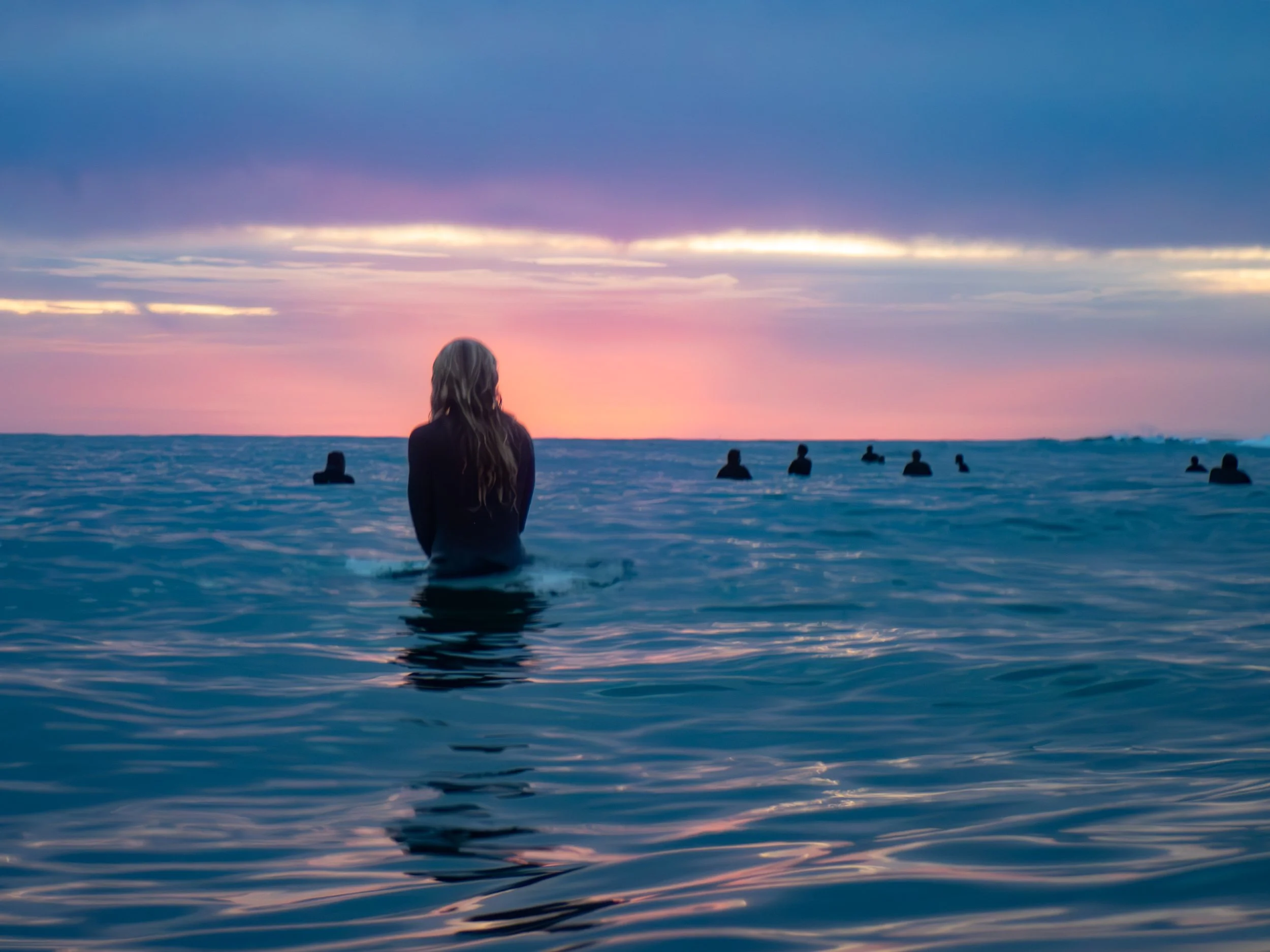 Woman standing in the ocean during sunset with several people in the water in the background.
