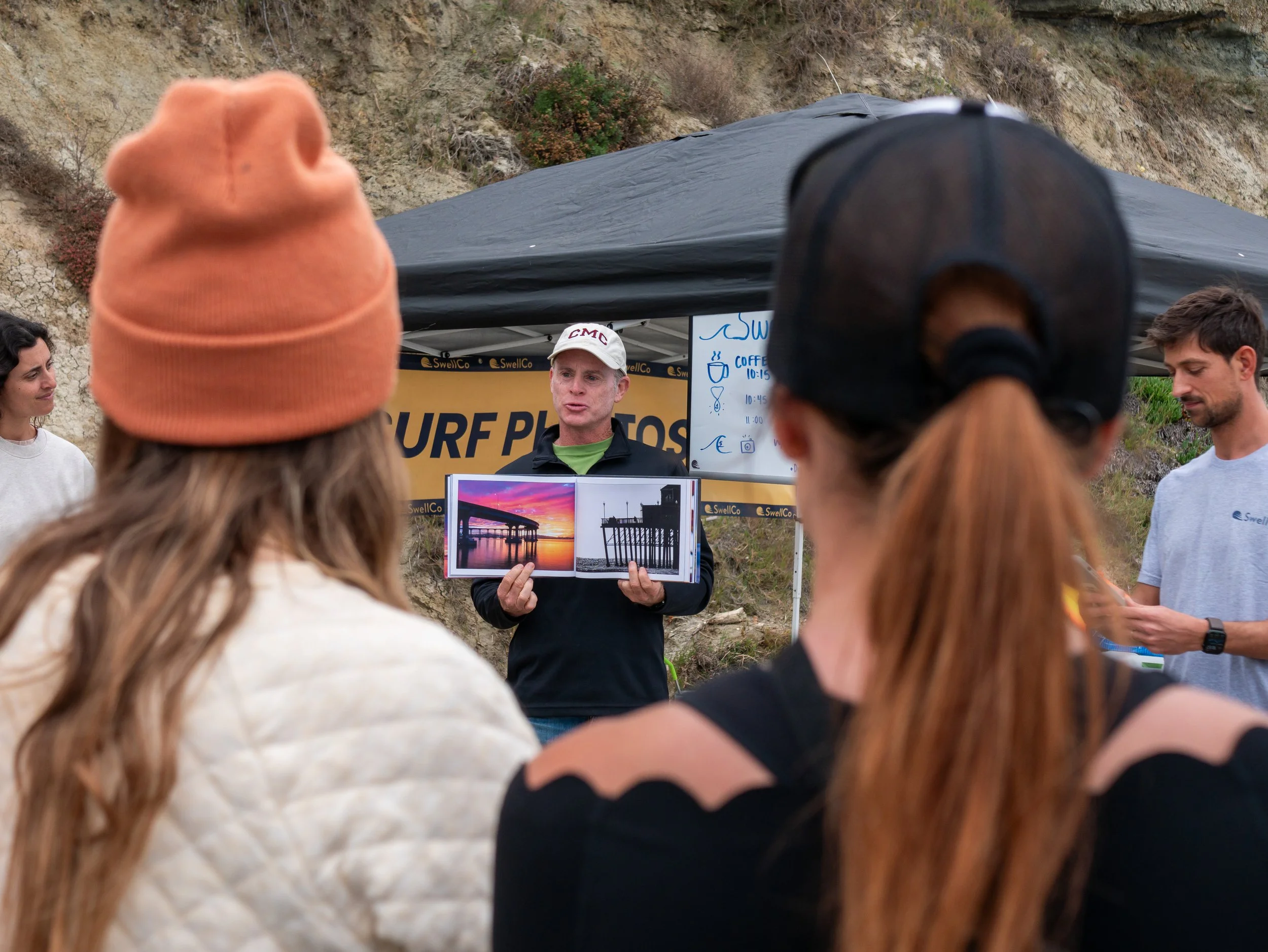 A man giving a presentation outdoors to a group of people, holding a photo album with sunset and pier images, under a black canopy with a sign that reads 'SURF PHOTOS' in the background.