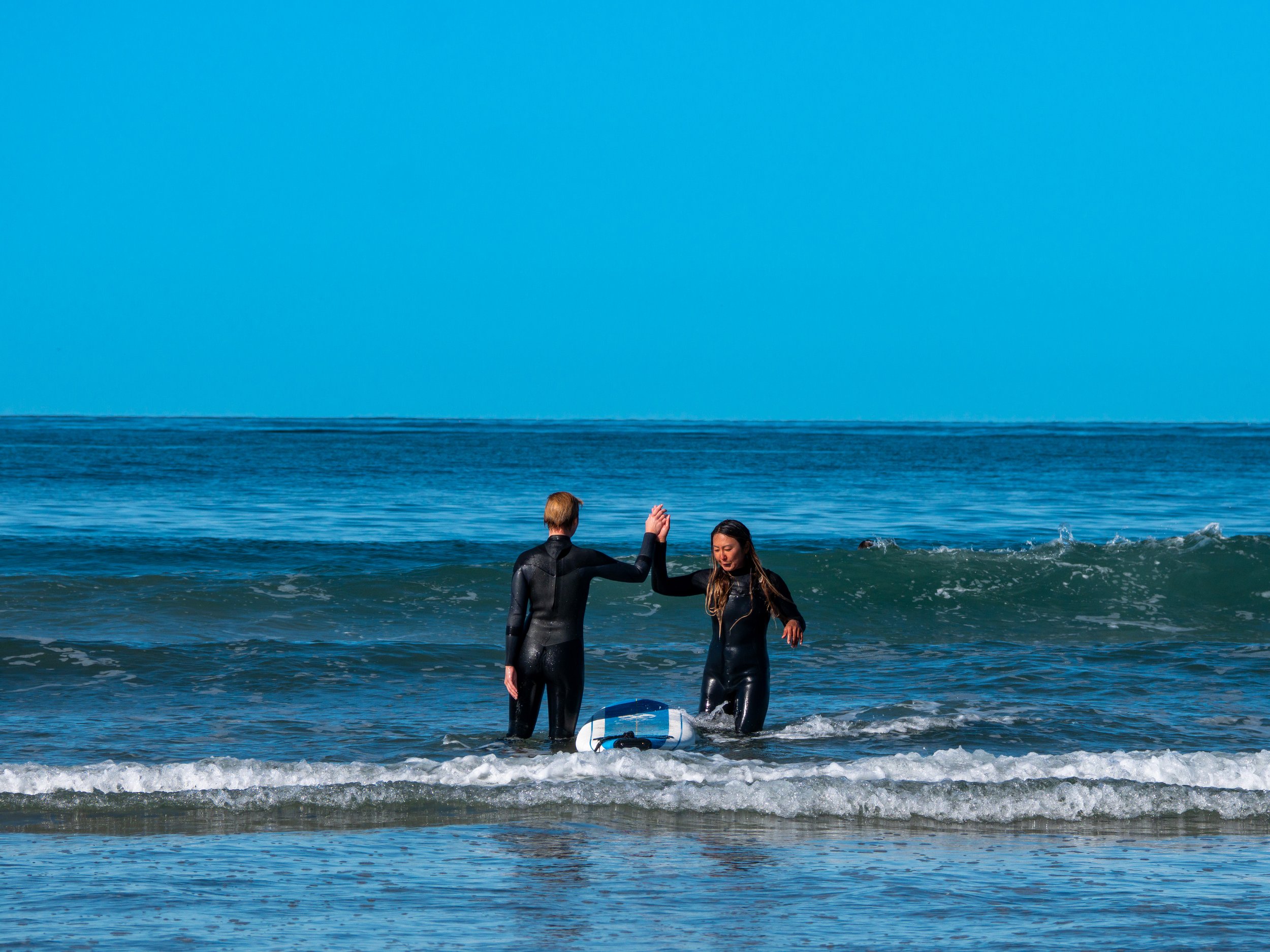 Two surfers in wetsuits giving each other a high-five in the ocean with a surfboard nearby.