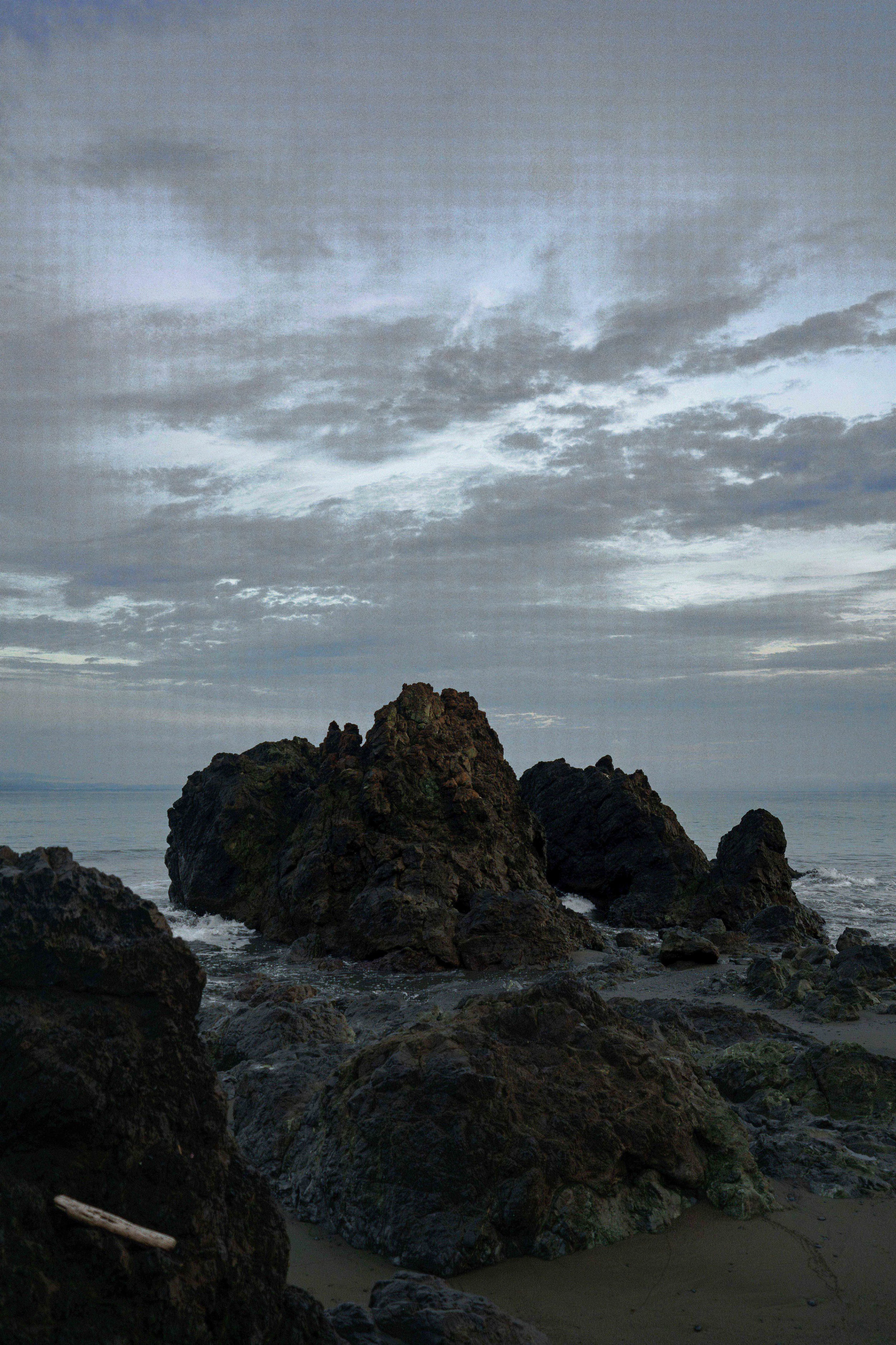 Overcast sky with scattered clouds over rocky shoreline and ocean, with large dark rocks in the foreground.