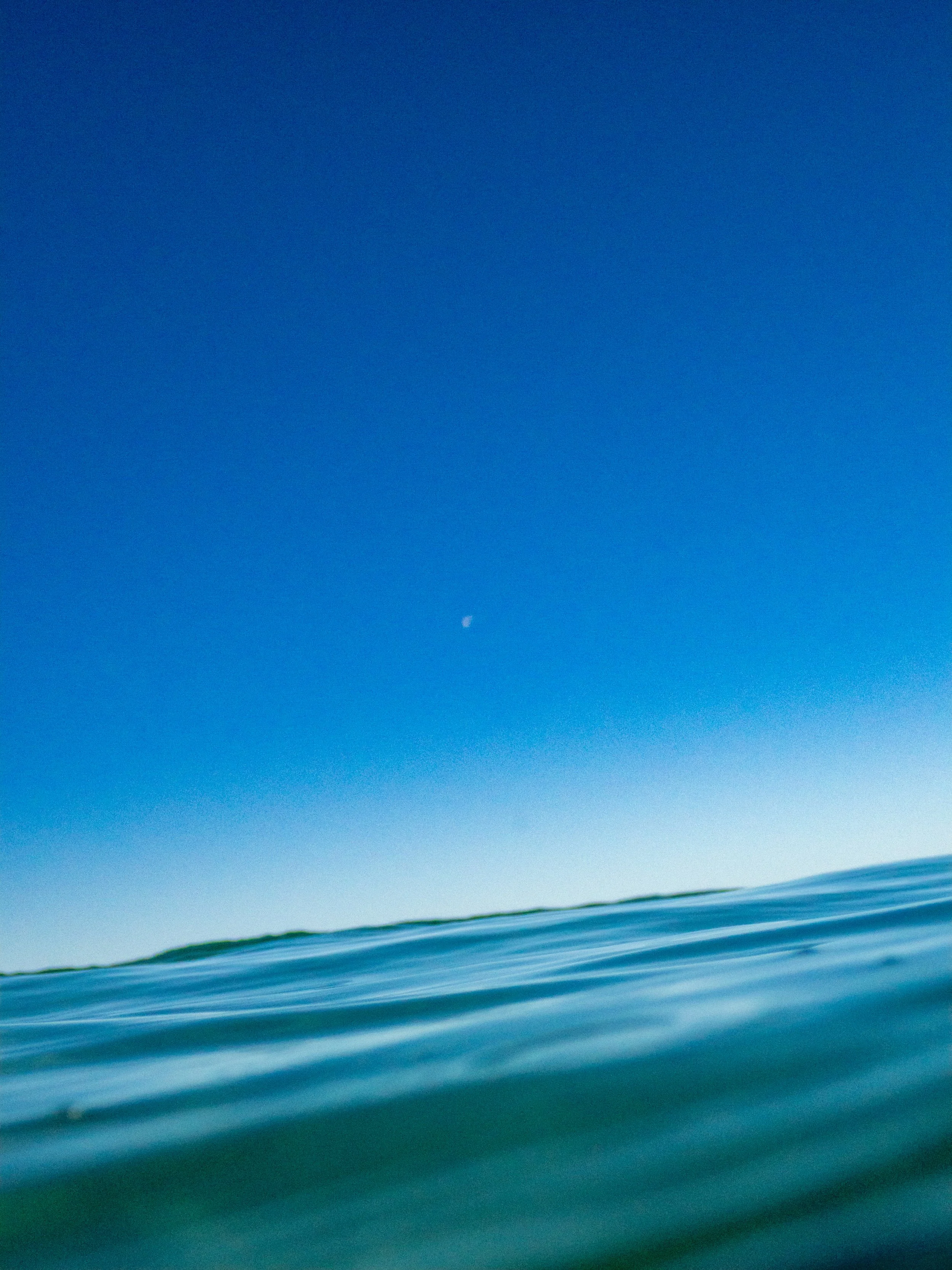 Ocean water with a clear blue sky and a small moon visible in the sky.