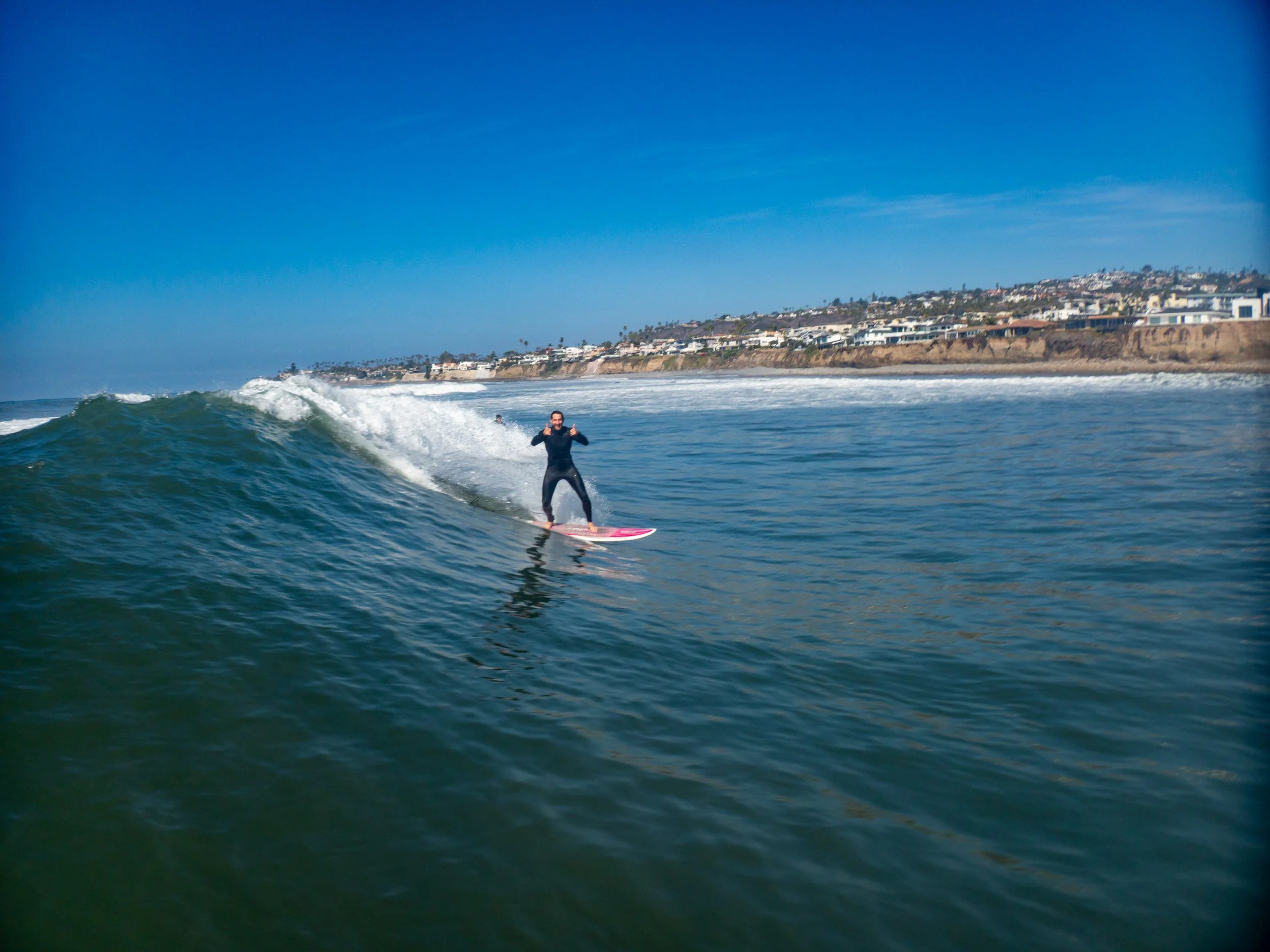 Person surfing on a wave off the coast with houses on a hill in the background and a clear blue sky.