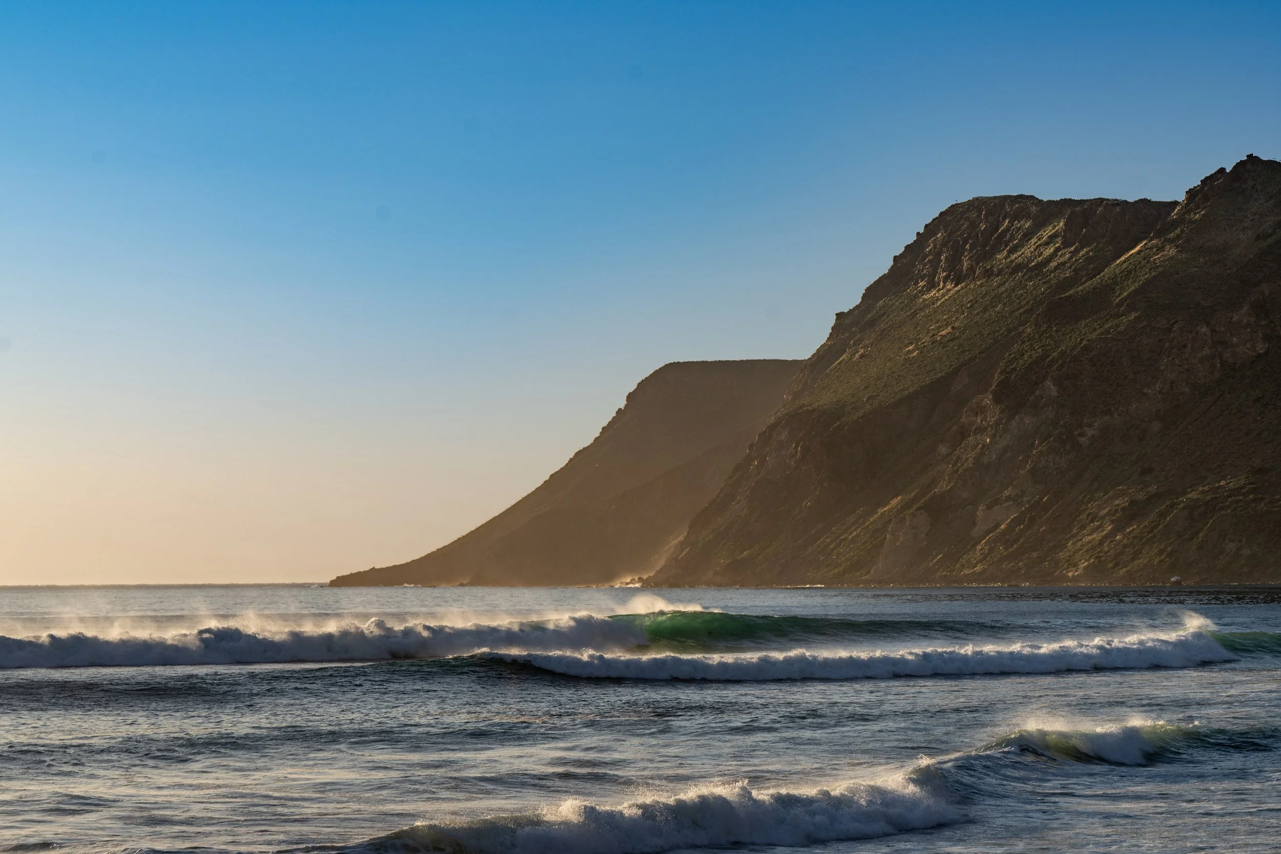 Ocean waves crashing near cliffs at sunset or sunrise, with a clear sky.