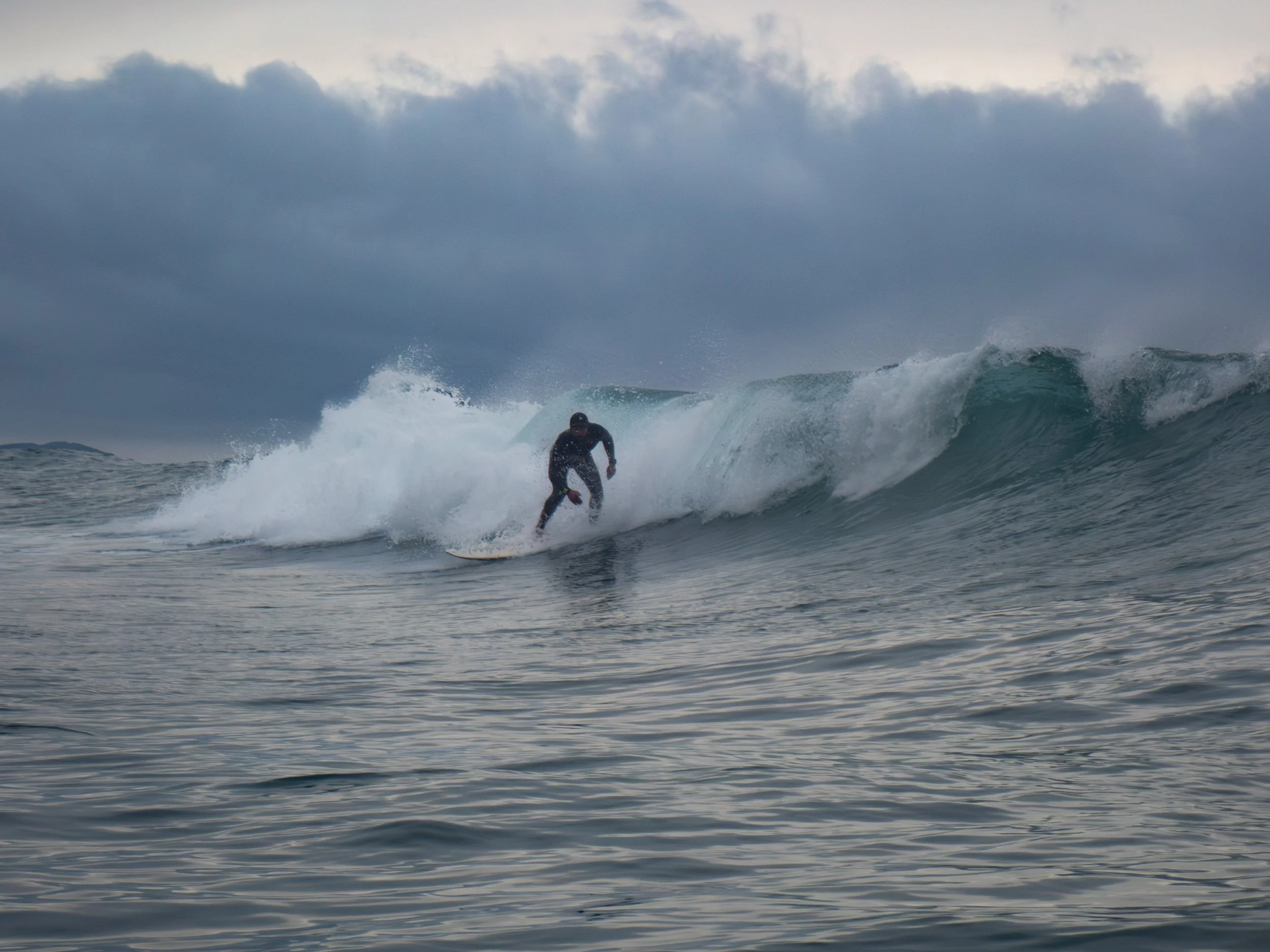 A person in black clothing surfing on a wave in the ocean under dark cloudy skies.