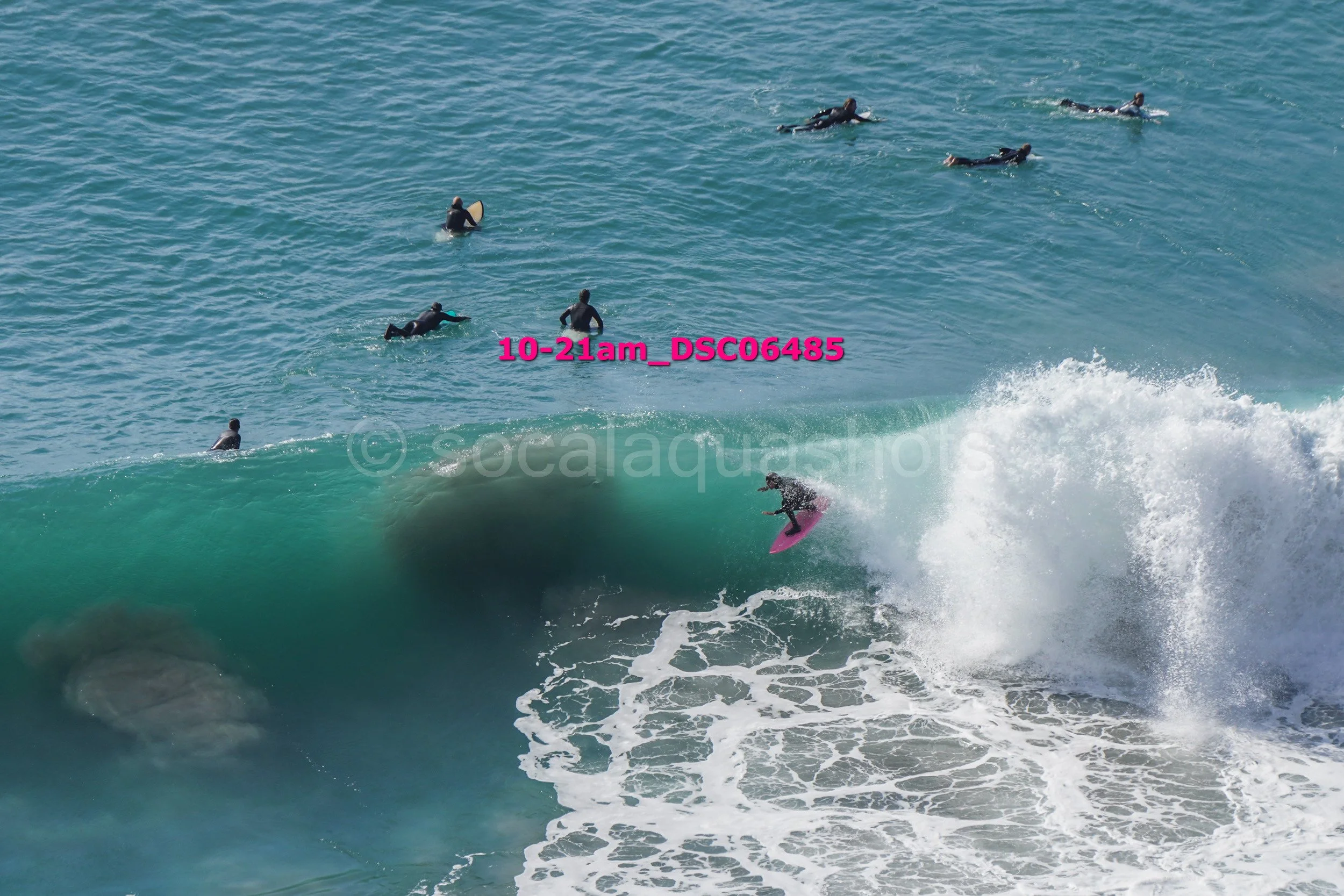 Surfer riding a large wave with several people waiting in the water nearby.