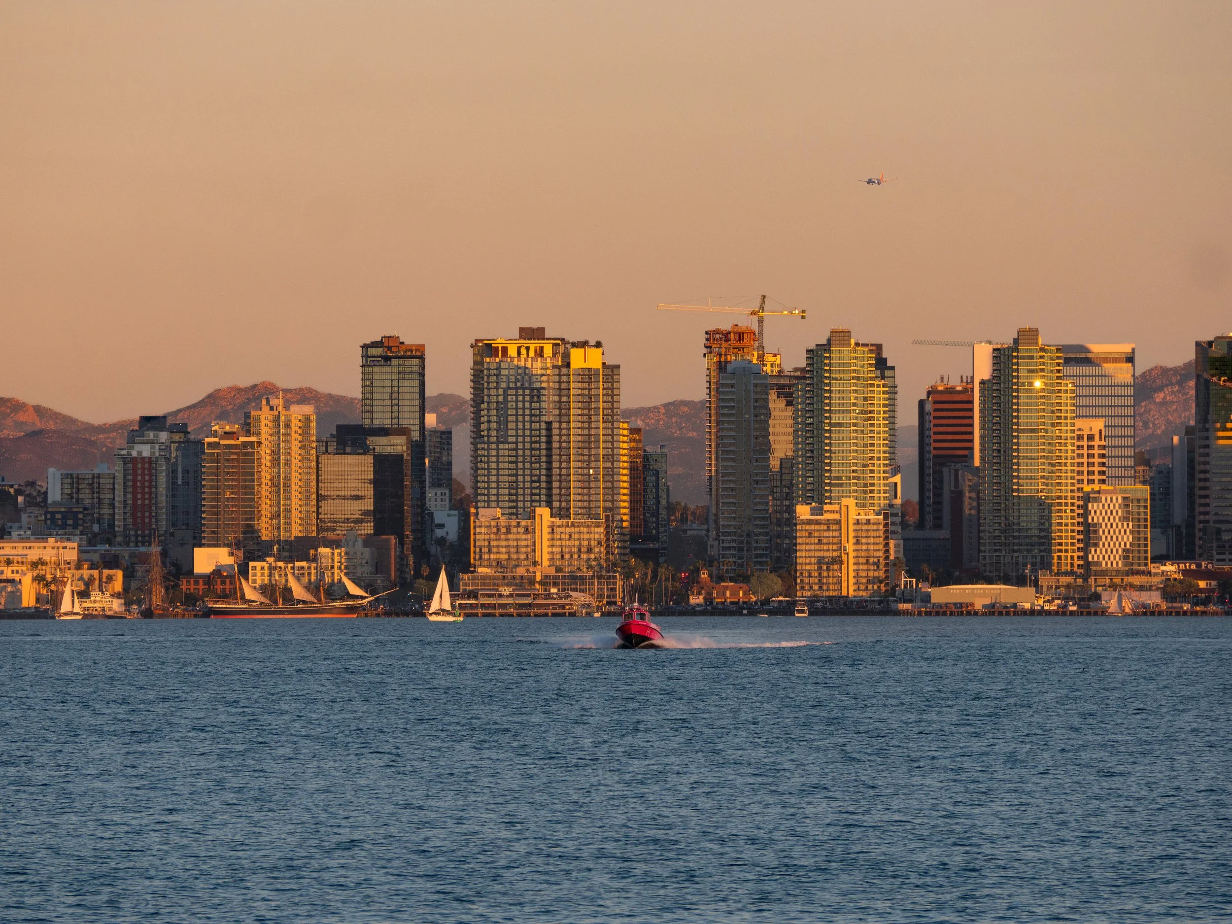 City skyline during sunset with high-rise buildings, a boat in the water, a helicopter flying above, and distant mountains in the background.