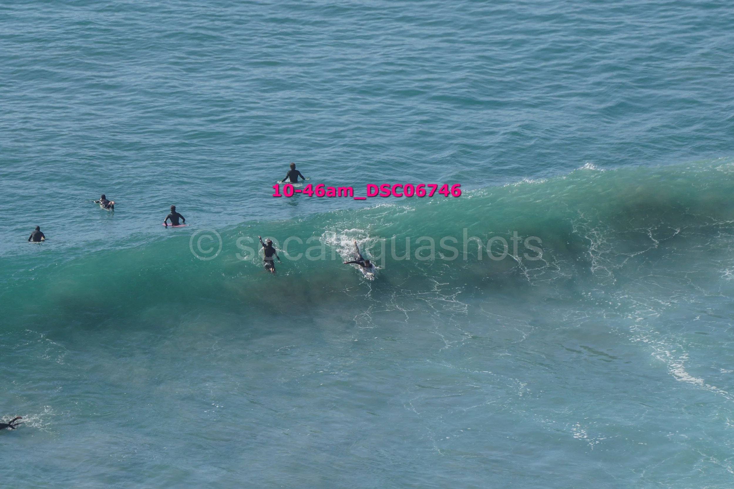 Multiple surfers waiting for waves in the ocean, some riding a large wave while others are paddling or waiting for their turn.