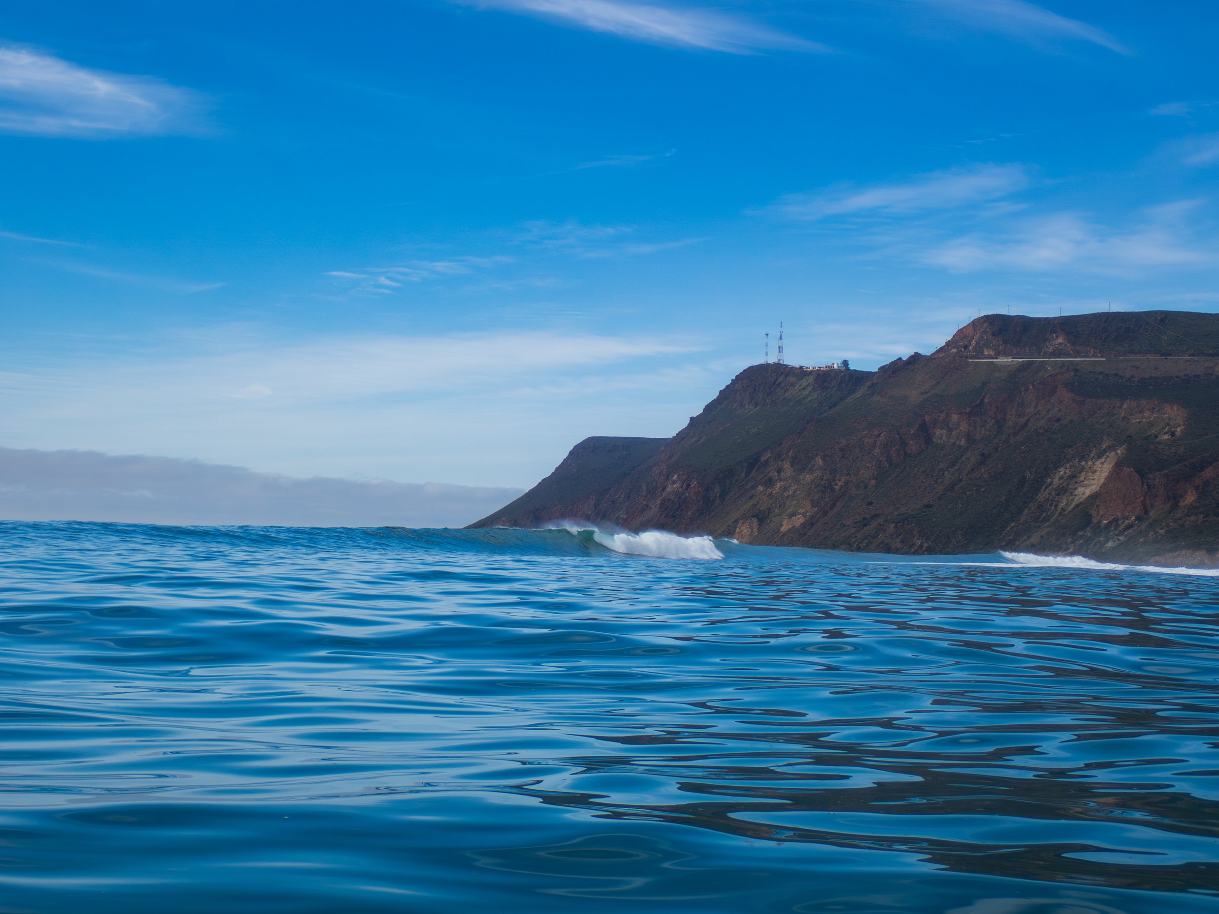 Ocean with small waves and a cliff in the background under a partly cloudy blue sky.