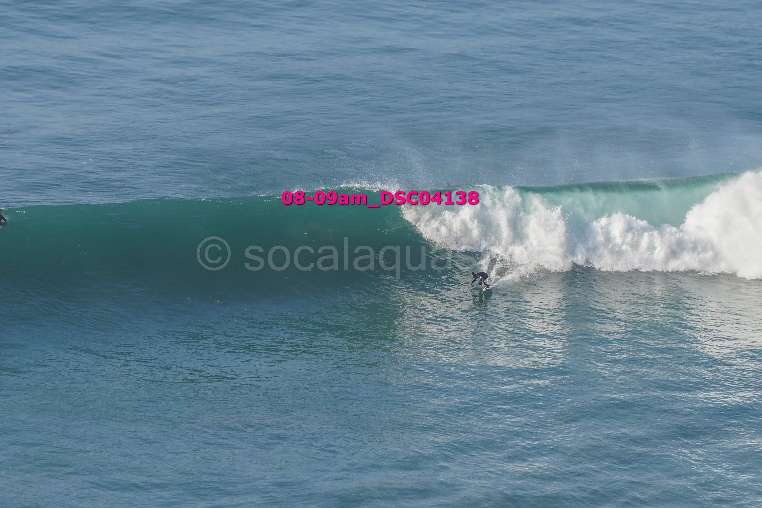 A person surfing on a small wave in the ocean with another surfer visible in the background.
