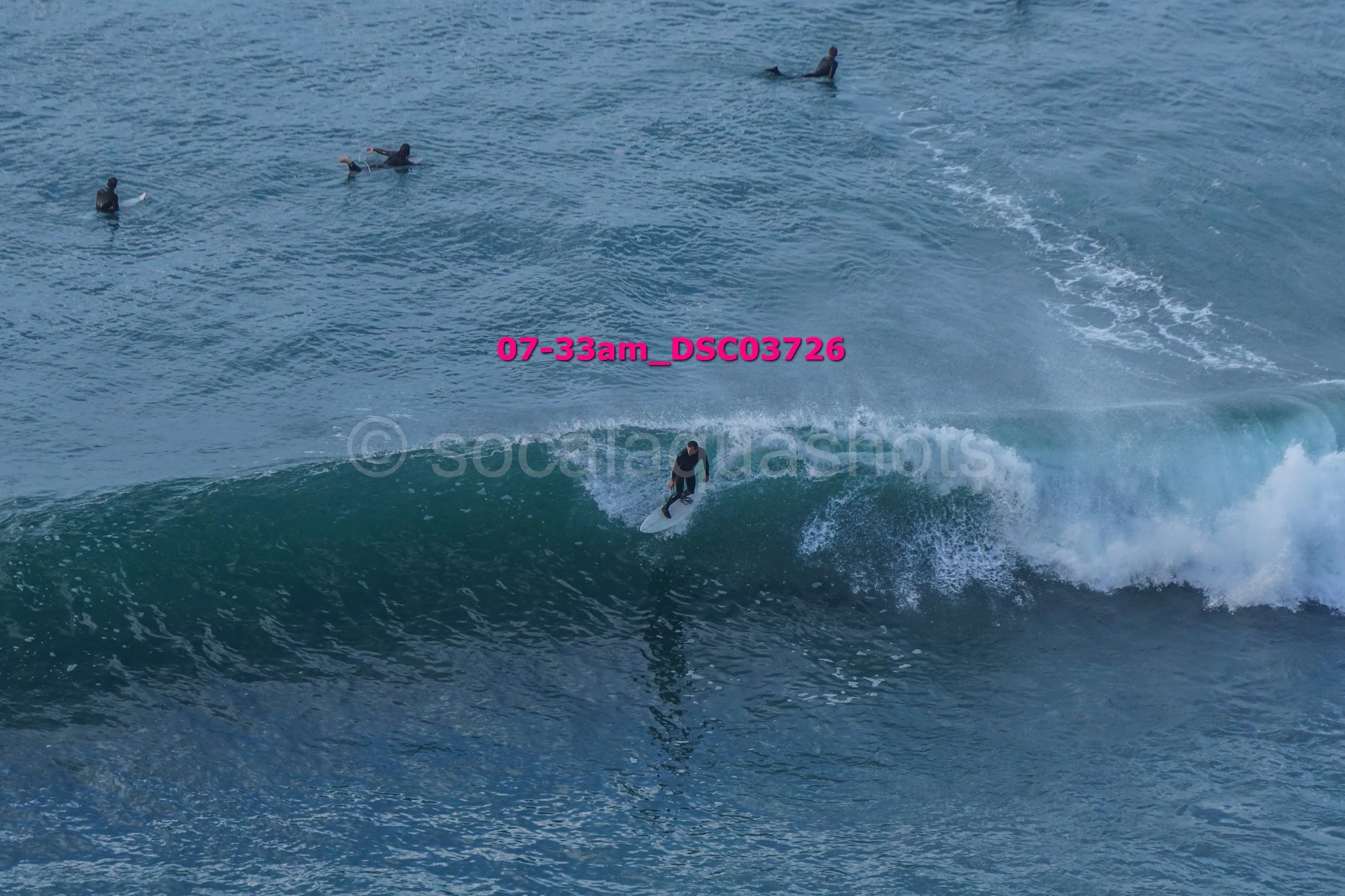 A surfer riding a large wave with four other surfers surfing in the background on the ocean.