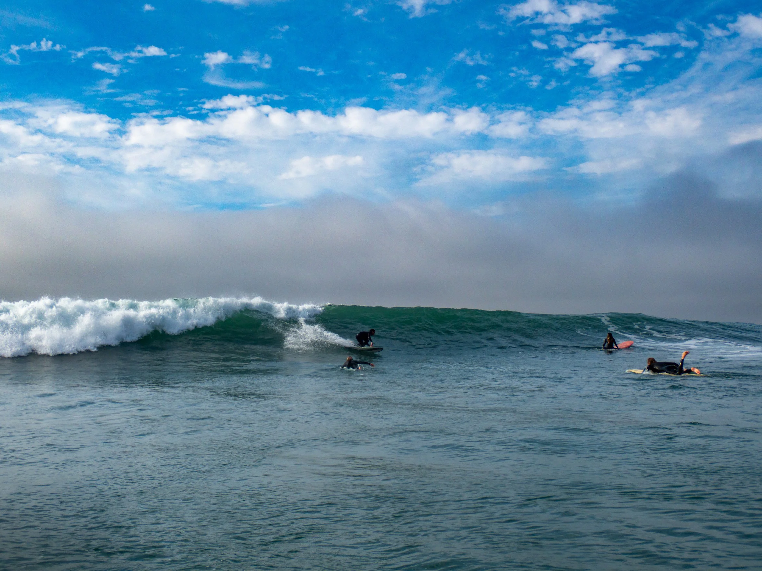 Four surfers in wetsuits riding and waiting for waves in the ocean under a partly cloudy sky.