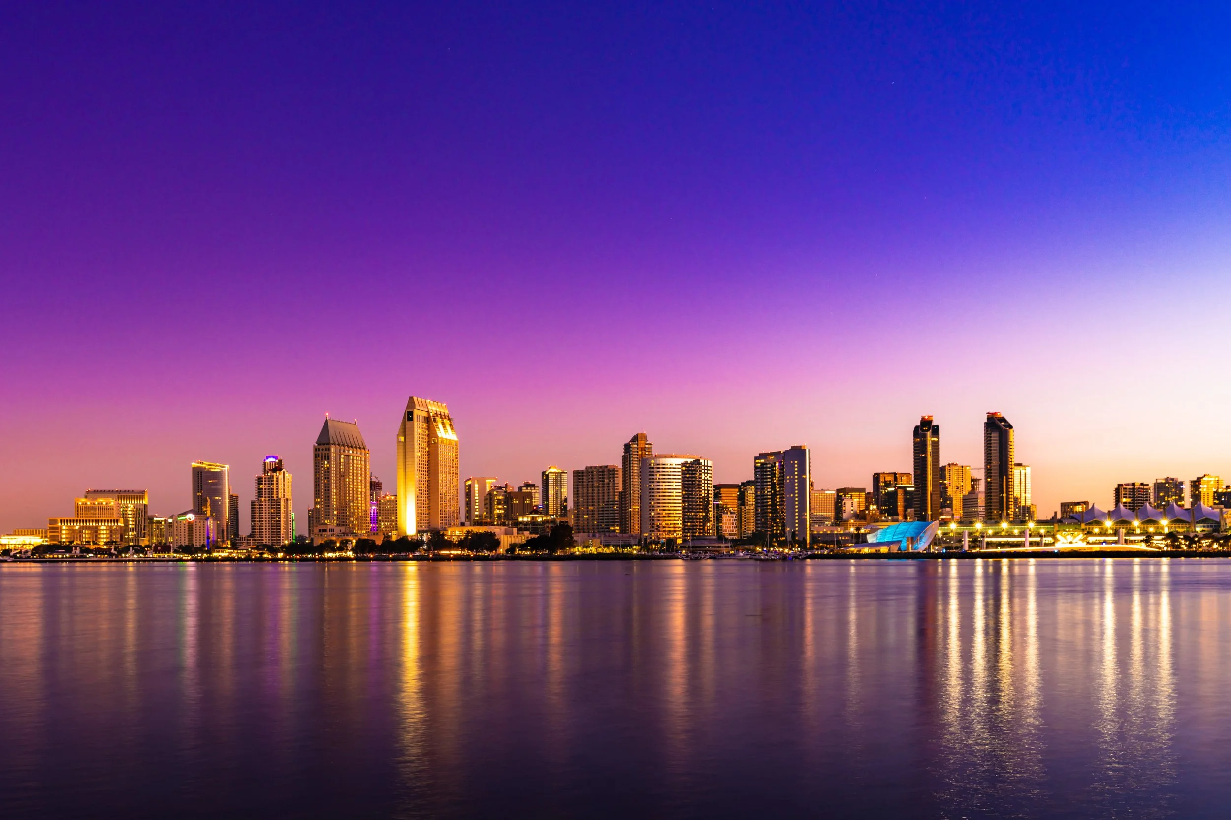 City skyline during sunset with tall buildings reflected in a body of water in the foreground.