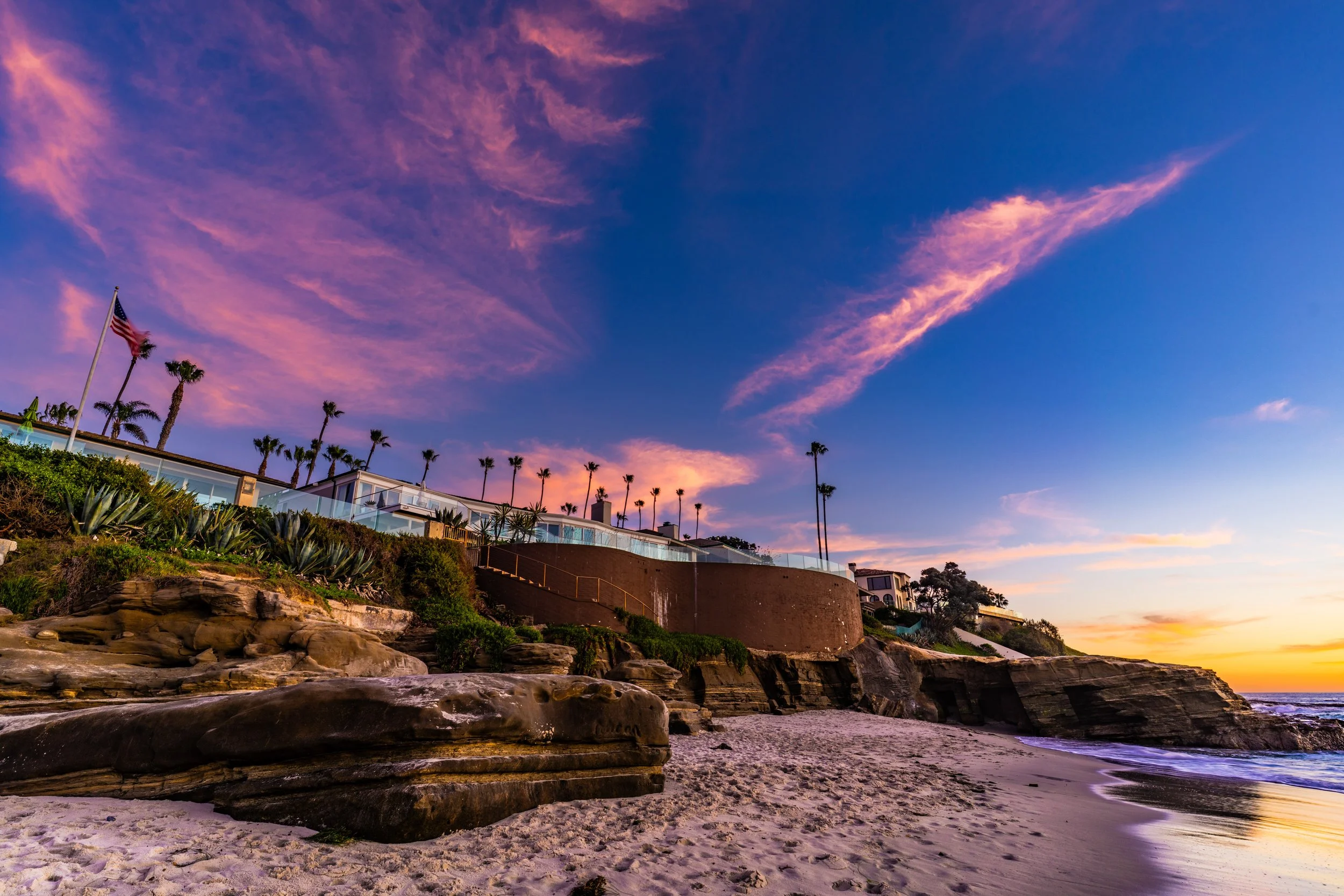 Sunset over a beach with rocky cliffs, sandy shore, and houses with palm trees on a hill.
