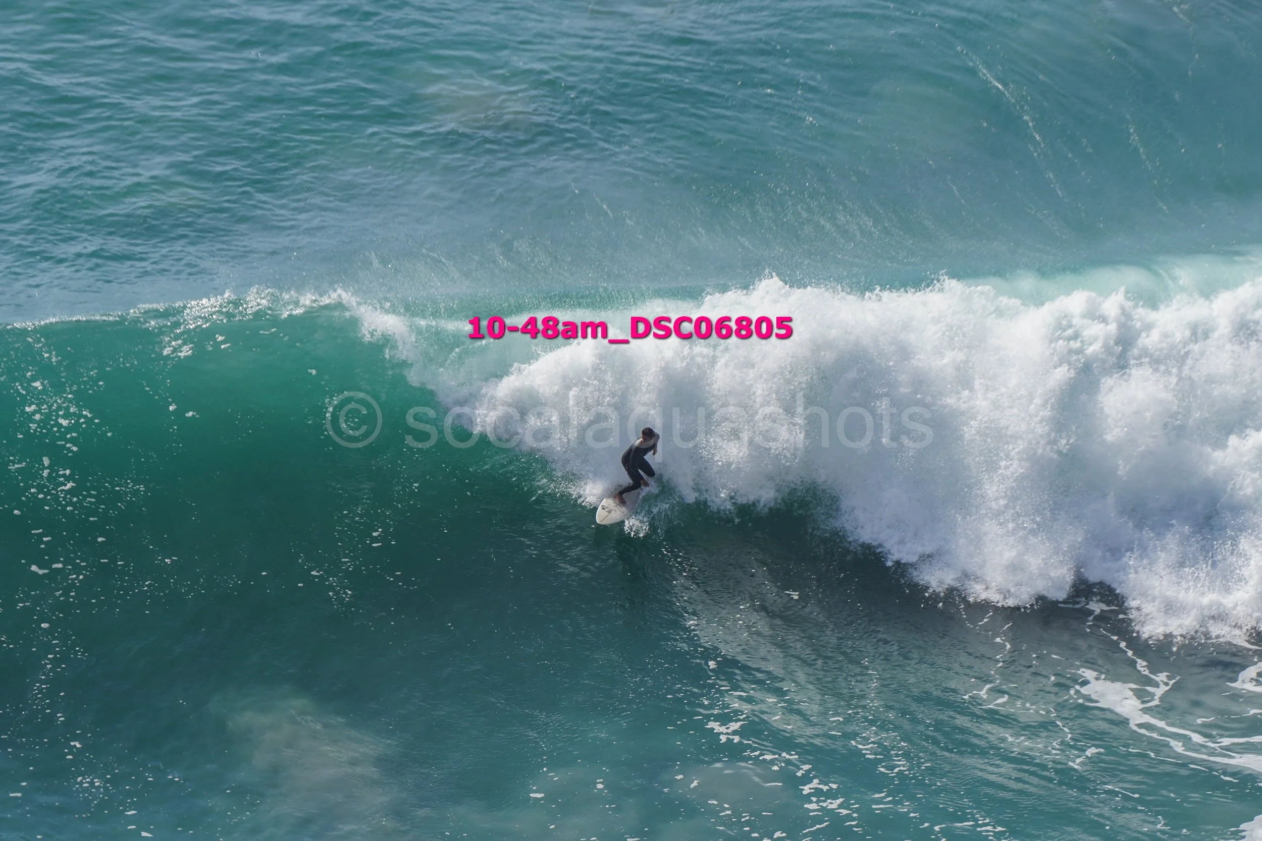 A person surfing on a large ocean wave with white foam and clear water, wearing a wetsuit.
