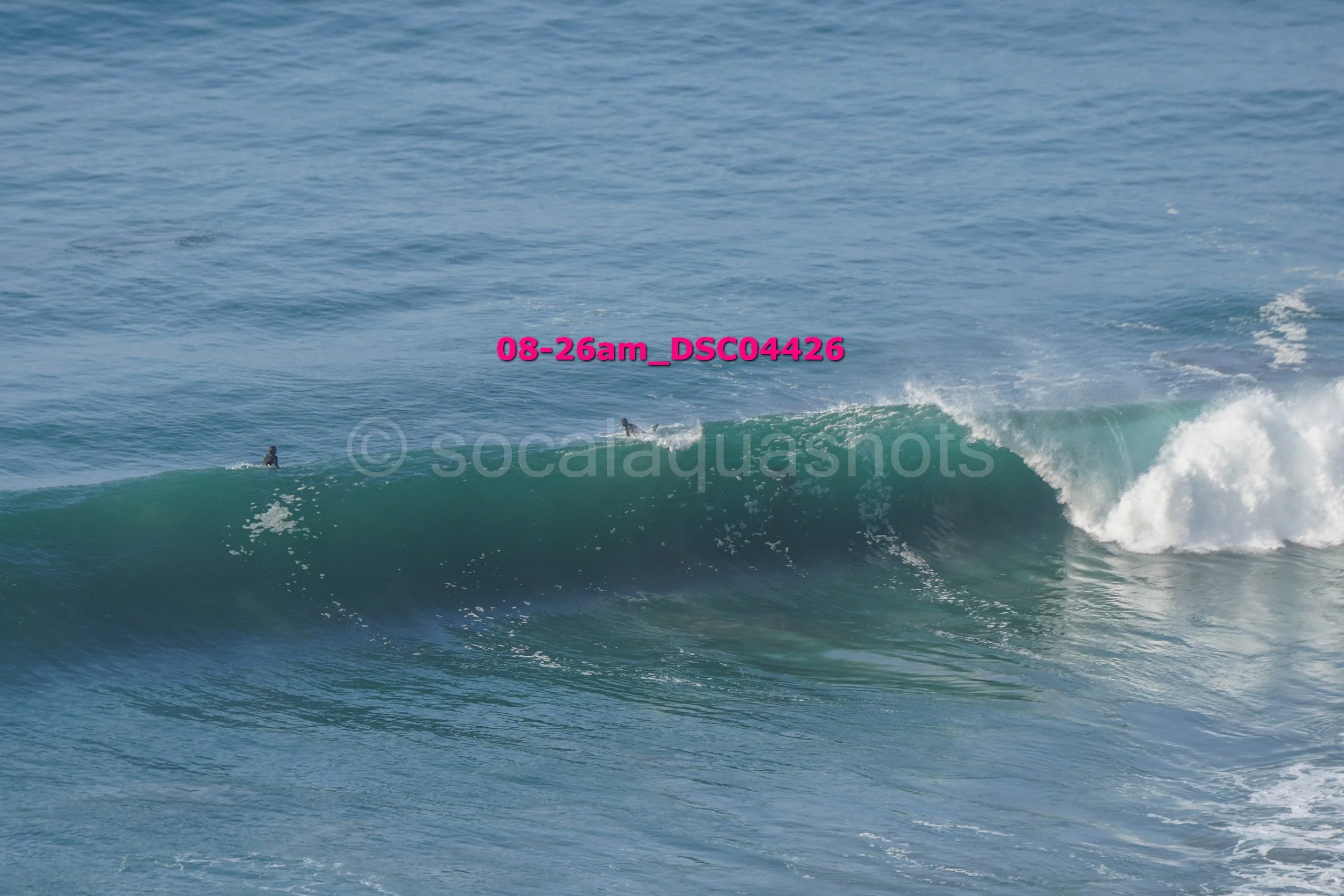 A large ocean wave with surfers in the water, one about to ride the wave, and the wave cresting with white foam.