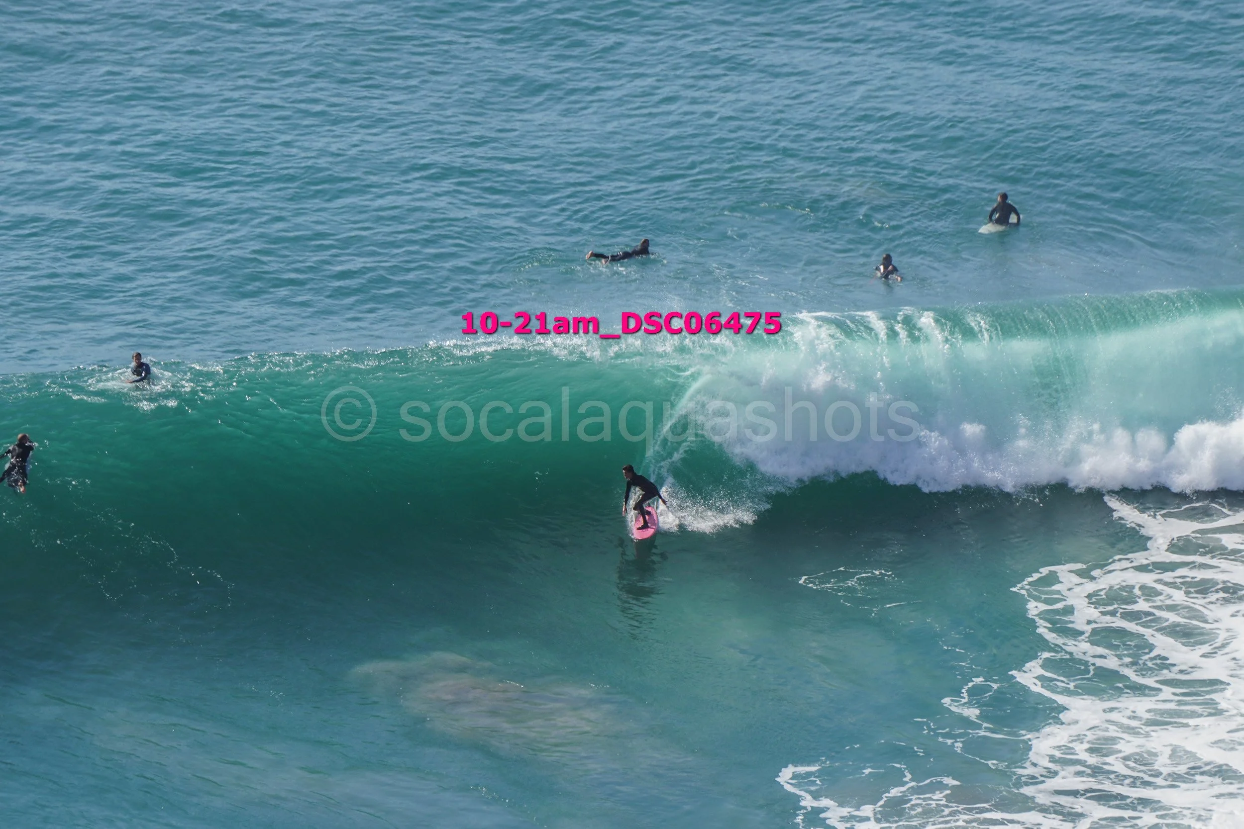Surfers riding and waiting for waves in the ocean, with one surfer on a pink surfboard riding inside a large wave.