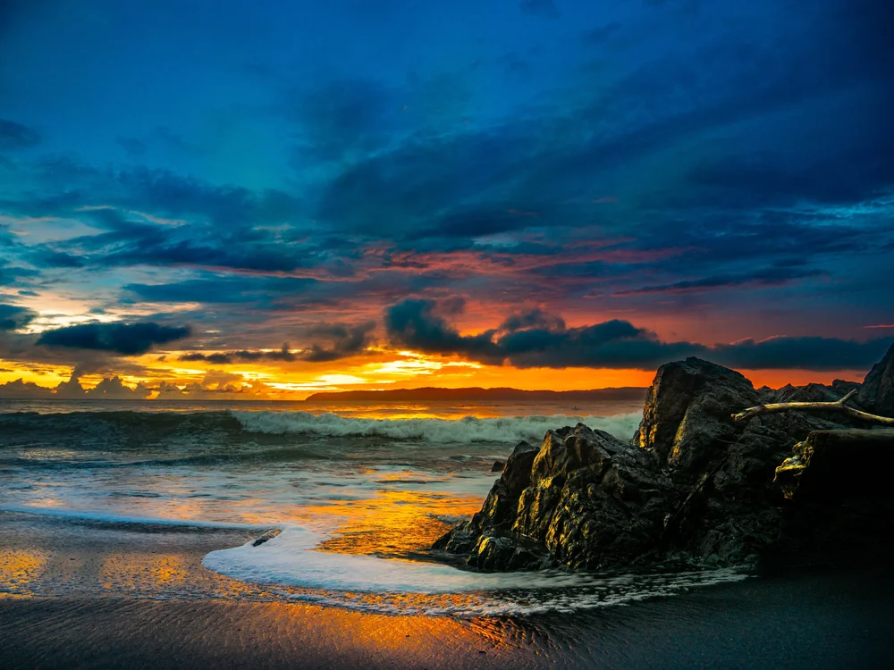 A dramatic Pavones sunset above breaking surf, dark clouds and shoreline rocks.