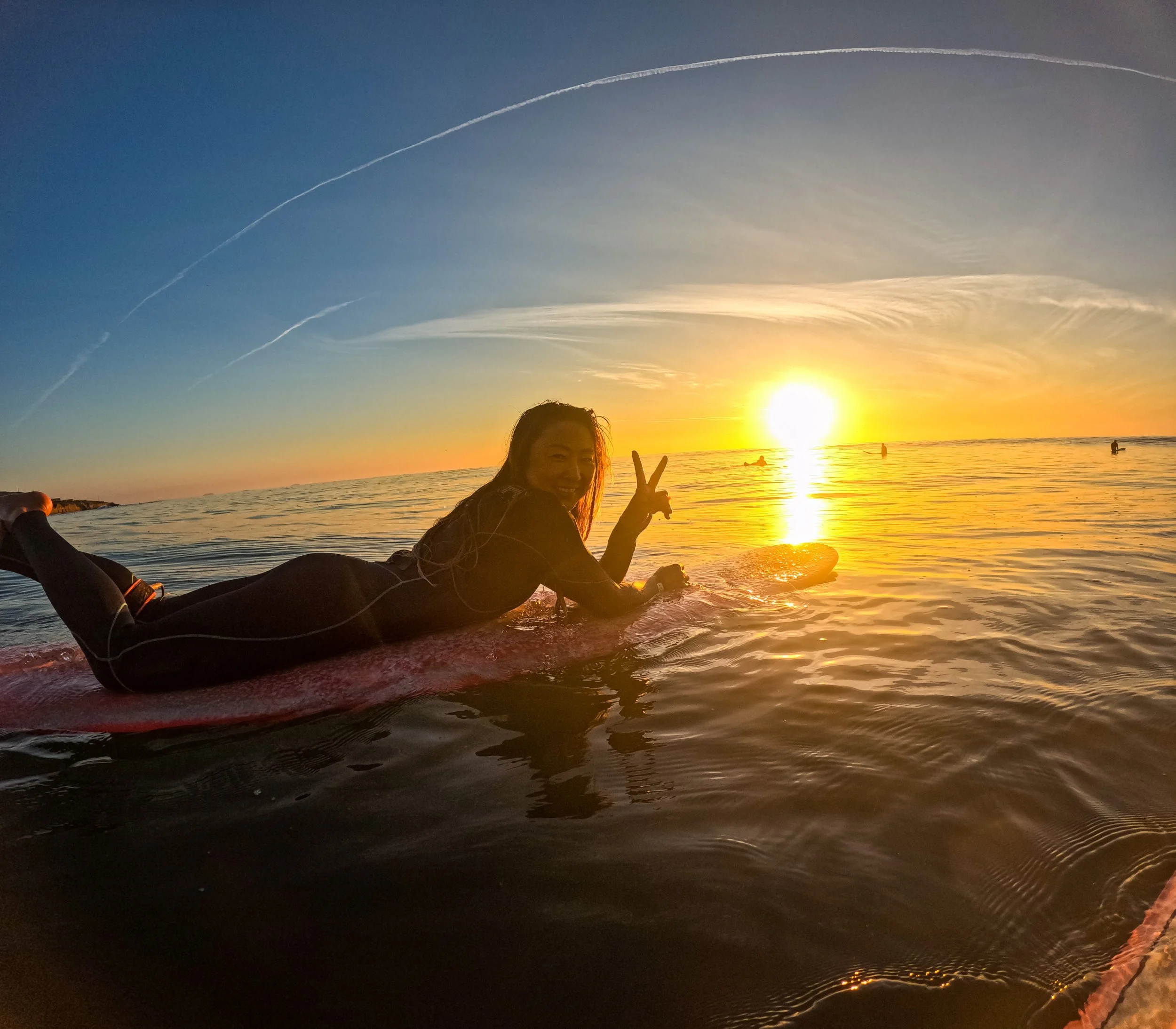 A woman in a wetsuit lying on a surfboard in the ocean during sunset, making a peace sign with her hand.